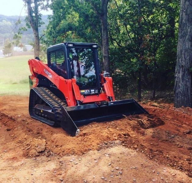 A small Kubota excavator on a dirt patch with trees and grass in the background.