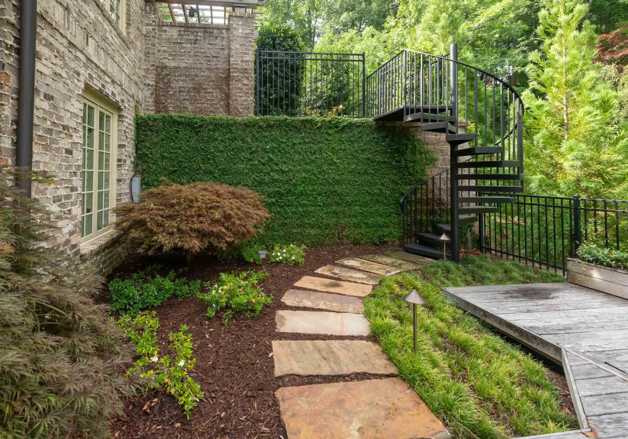 A backyard garden with a stone pathway, lush plants, and a black metal spiral staircase against a brick wall and green trees.