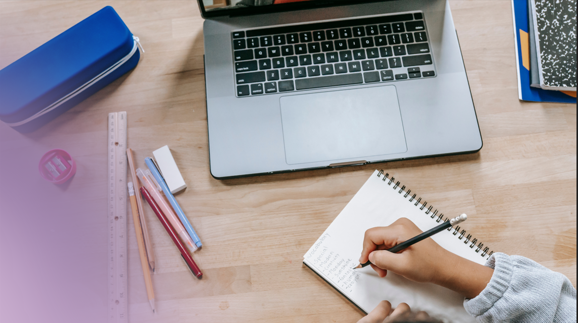 Mesa de estudo com laptop, cadernos, canetas, régua, fita adesiva, estojo azul, e uma pessoa escrevendo em um caderno