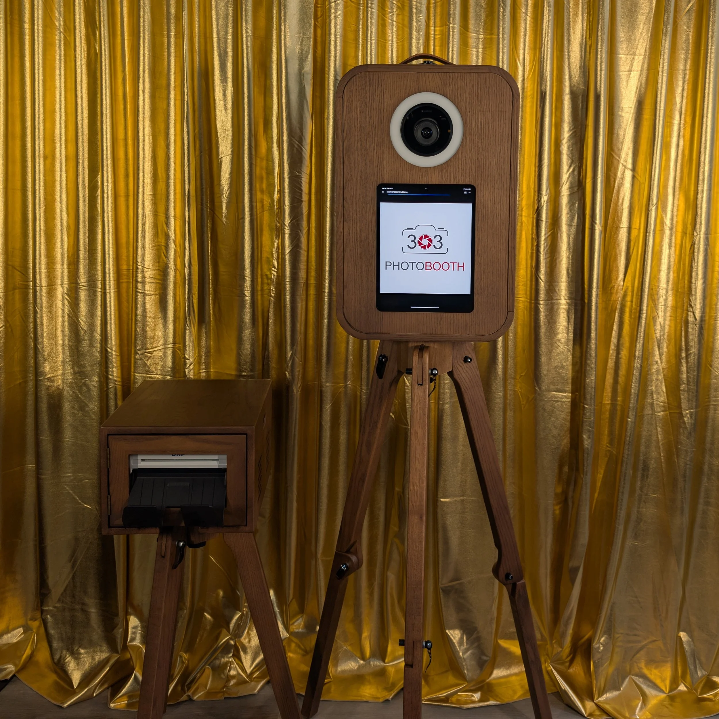 Photo booth with a camera on a tripod, a screen displaying the logo '303 PHOTOBOOTH', and a smaller device on a stand, set against a gold curtain background.