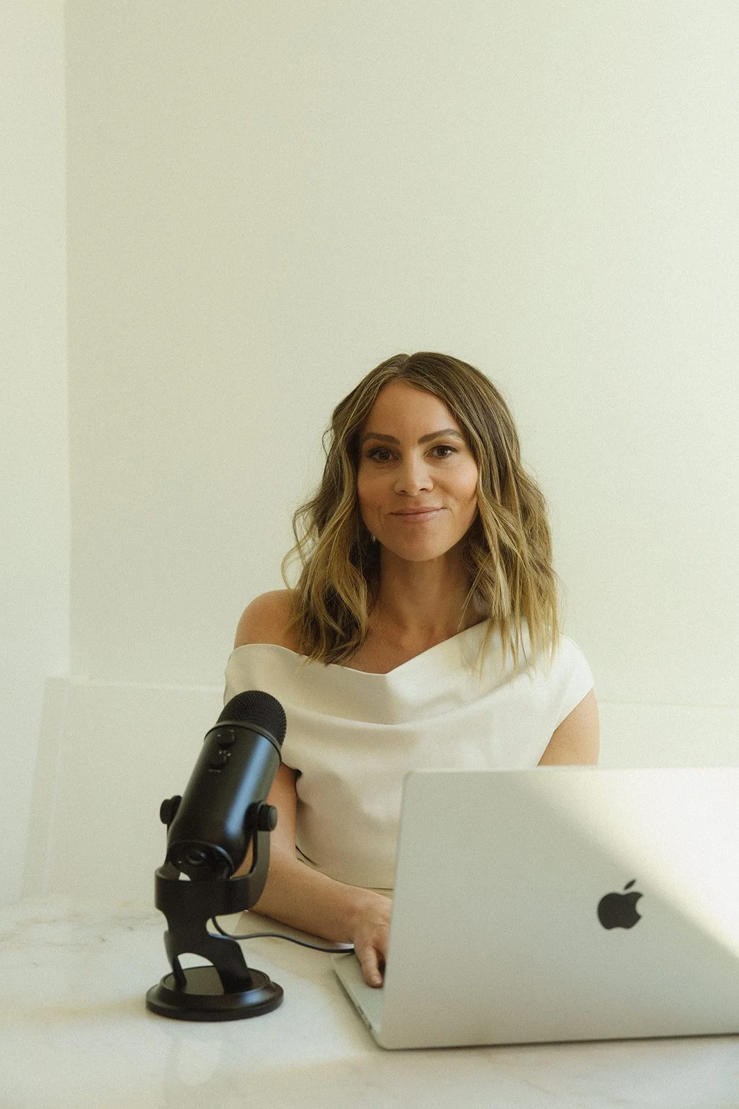 A woman sitting at a desk with a laptop and a microphone, in a white room.