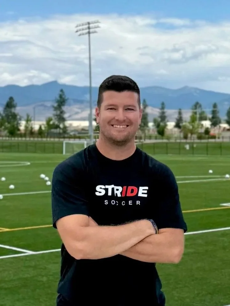 Man standing on a soccer field with arms crossed, smiling, wearing a black T-shirt that says 'STRIDE SOCCER,' with a goalpost, trees, mountains, and cloudy sky in the background.