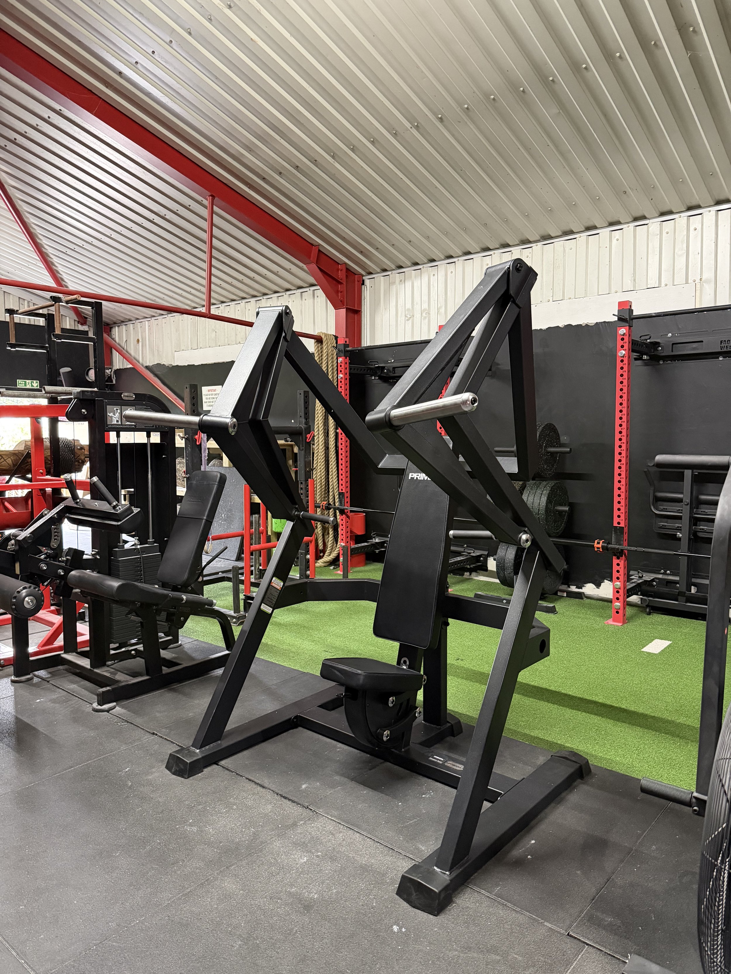 Indoor gym with strength training equipment, including a leg press machine, weight rack, and climbing ropes, set on a black rubber floor with a green artificial turf section. The ceiling is made of corrugated metal panels.