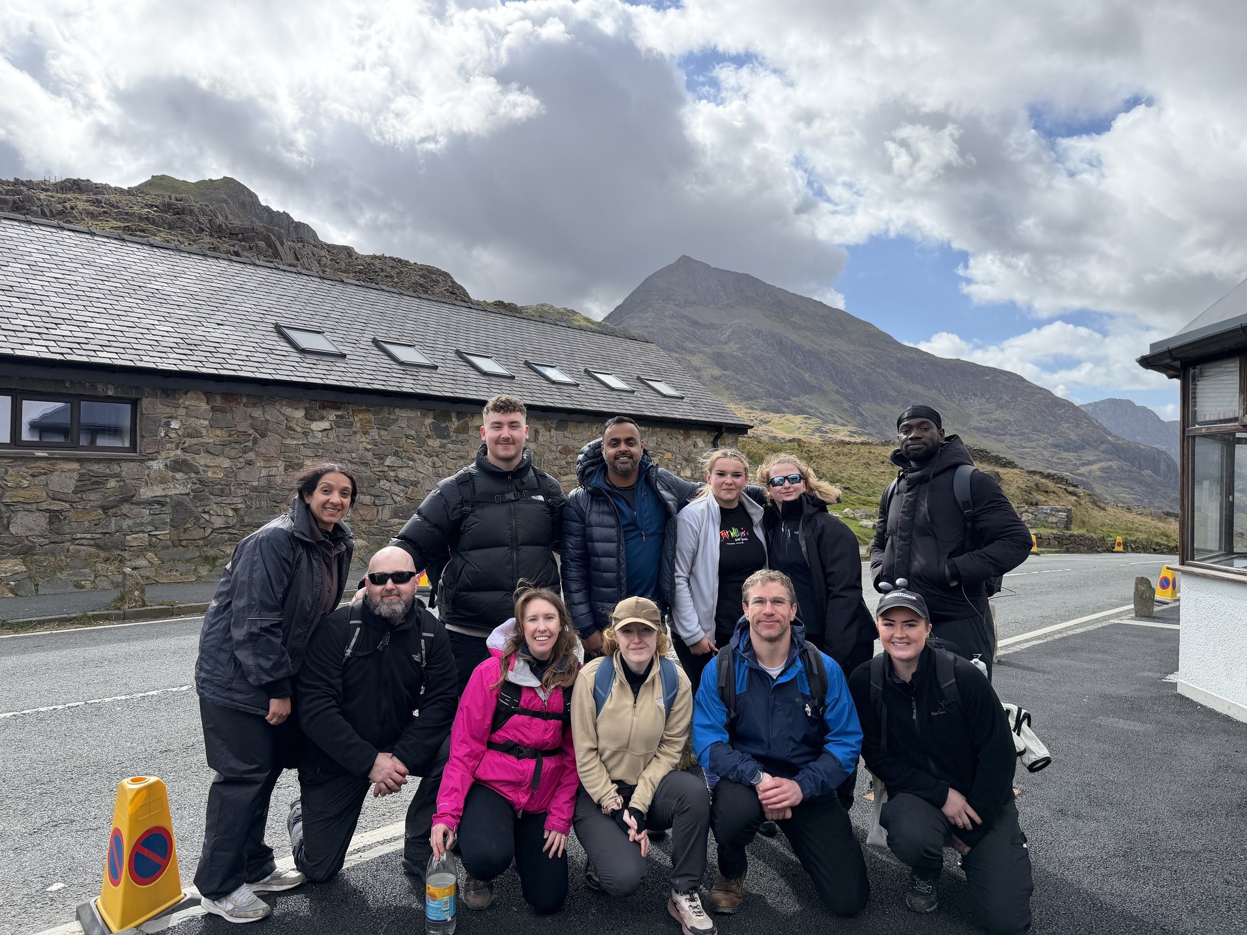 Group of people posing outdoors with mountains and partly cloudy sky in the background.