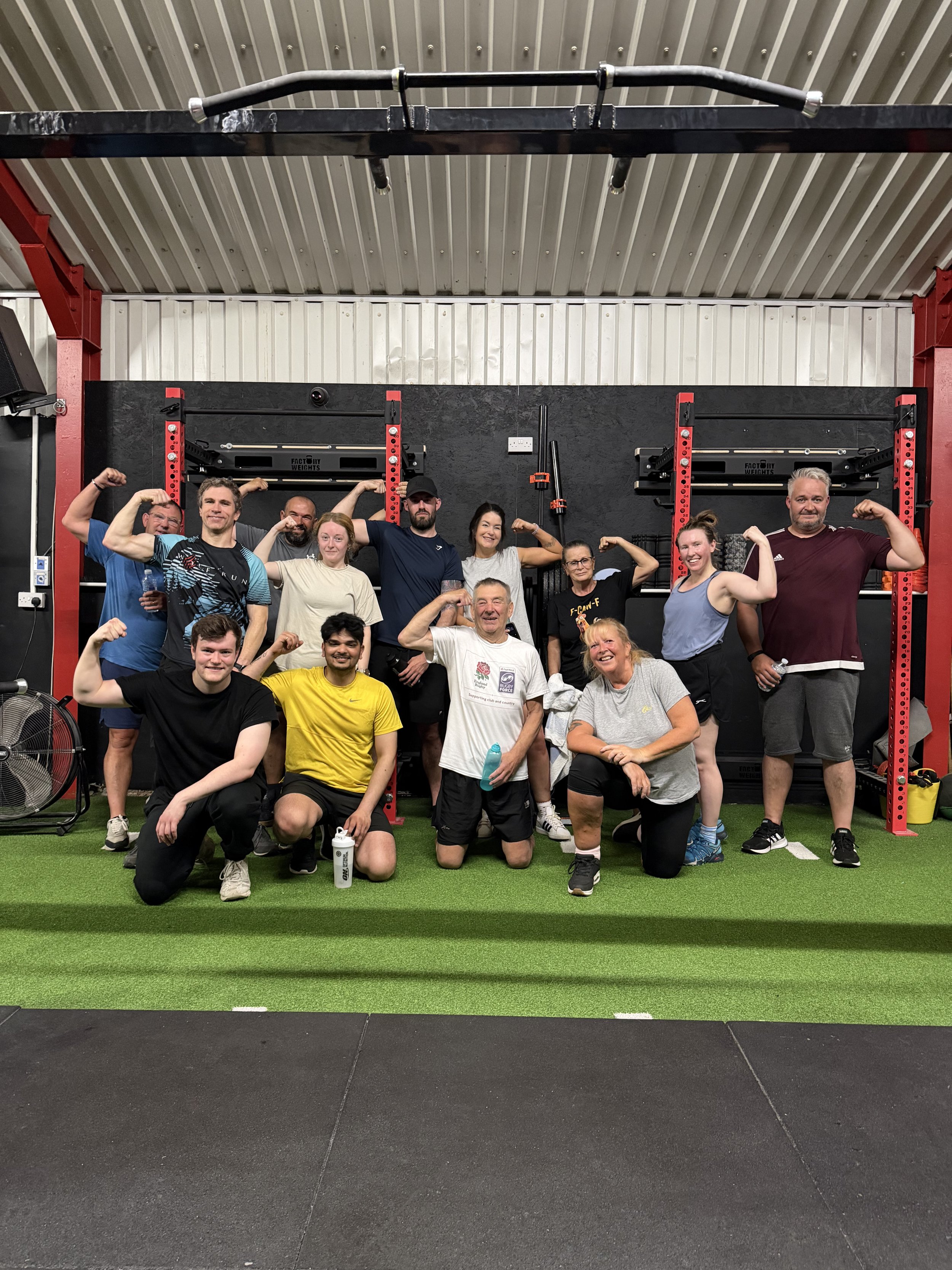 Group of people flexing their muscles in a gym, with weightlifting equipment and a black wall in the background.