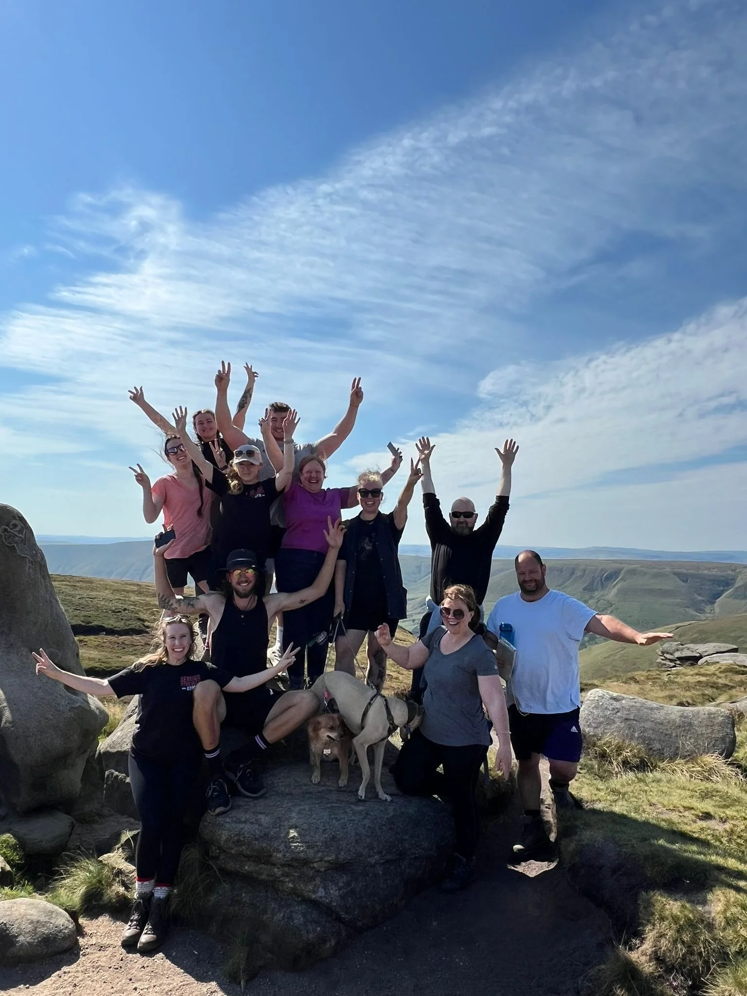 A group of people outdoors on a rocky landscape under a partly cloudy sky, smiling and making peace signs, with a dog among them.