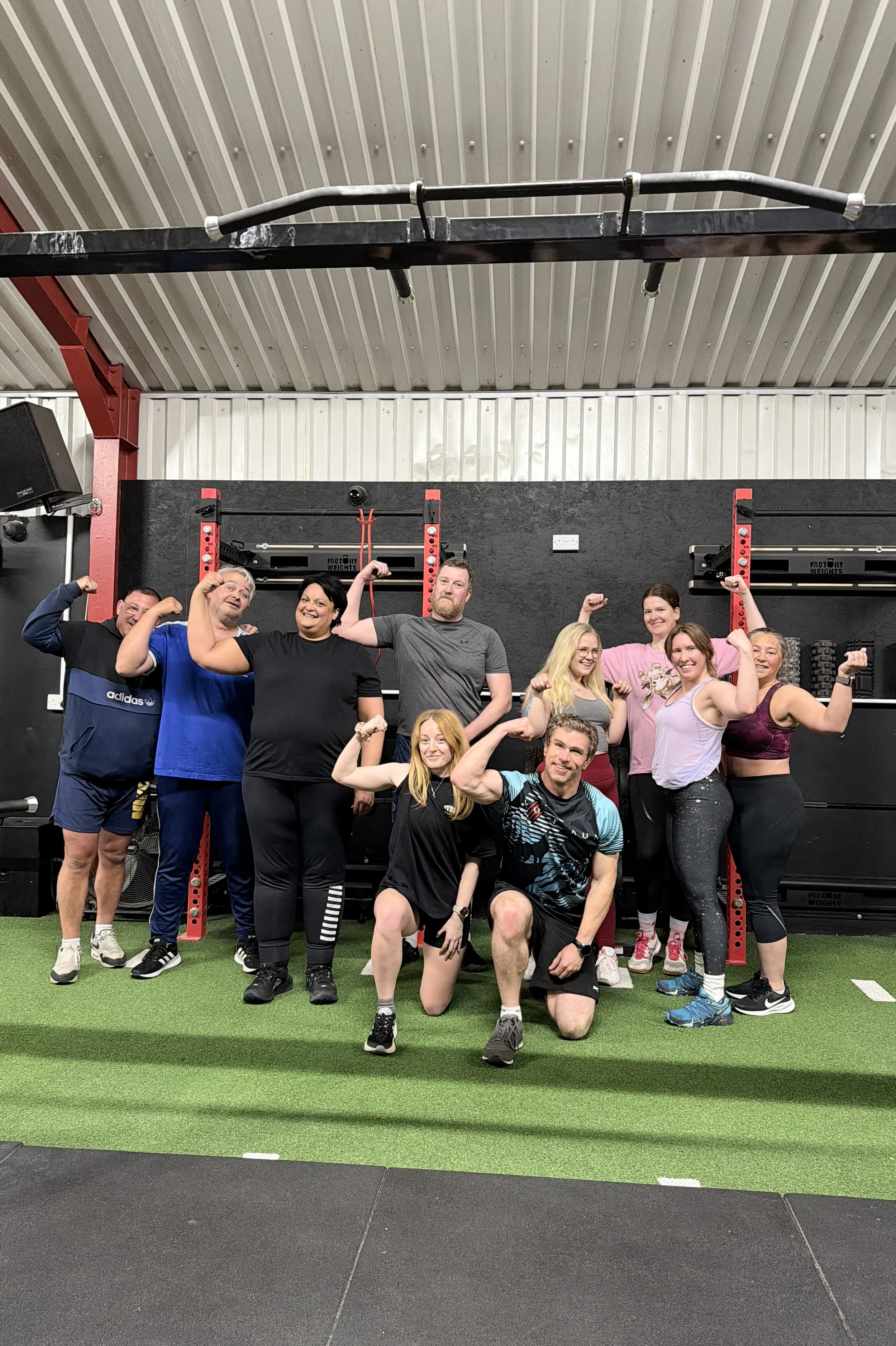 A group of ten people in a gym posing with their muscles flexed. They are standing on green and black gym flooring with fitness equipment and black wall panels behind them.