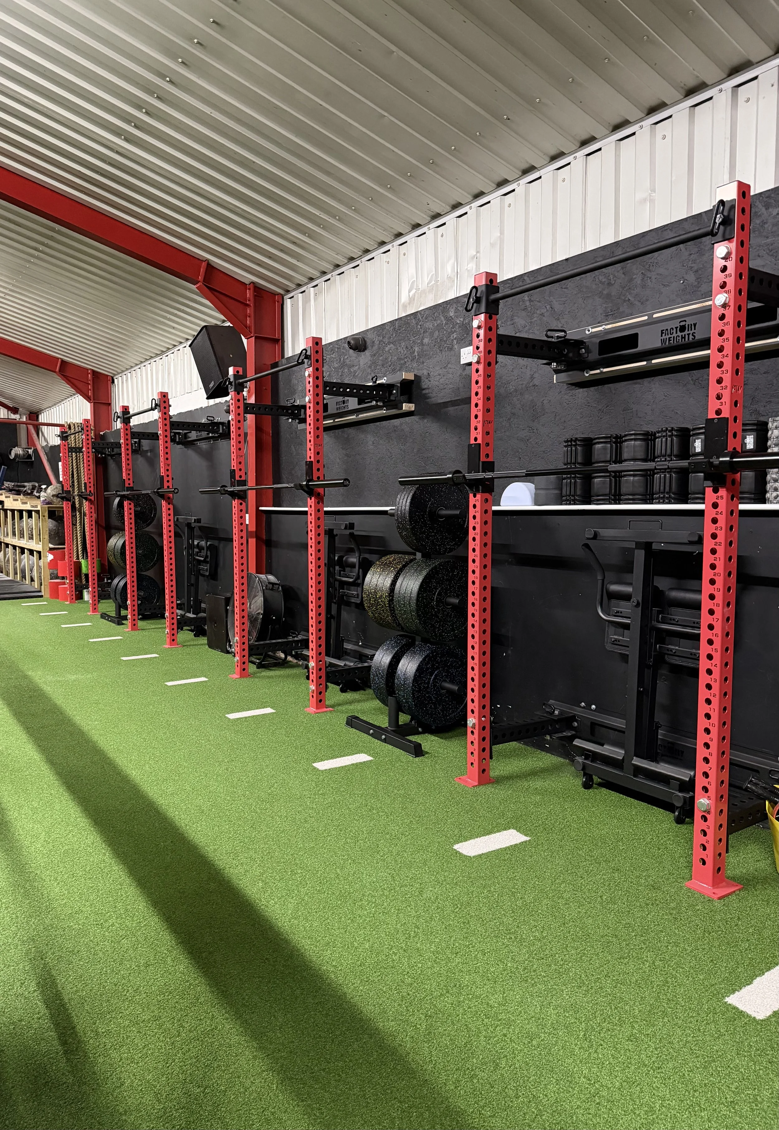Inside a gym with green turf flooring, red and black workout racks with weight plates, barbells, and resistance bands, against a black wall with white lines and a white ceiling with metal beams.