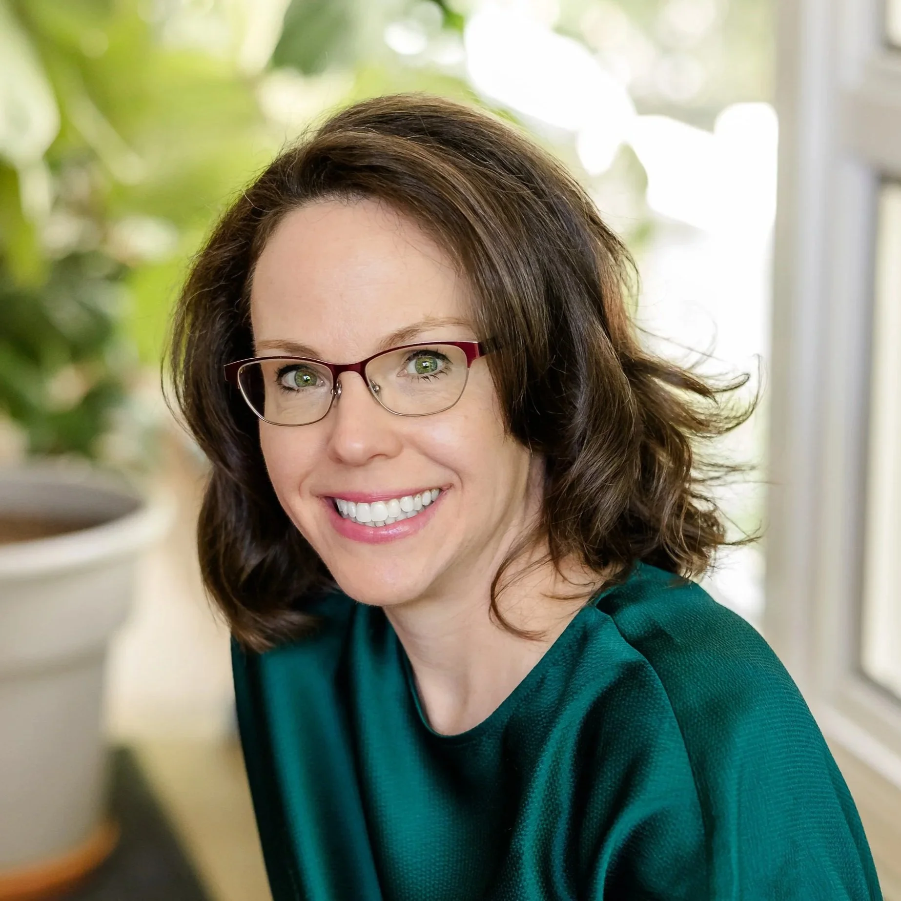 Close-up of a woman with brunette hair and glasses smiling, wearing a teal top, with a blurred background of greenery and a window.