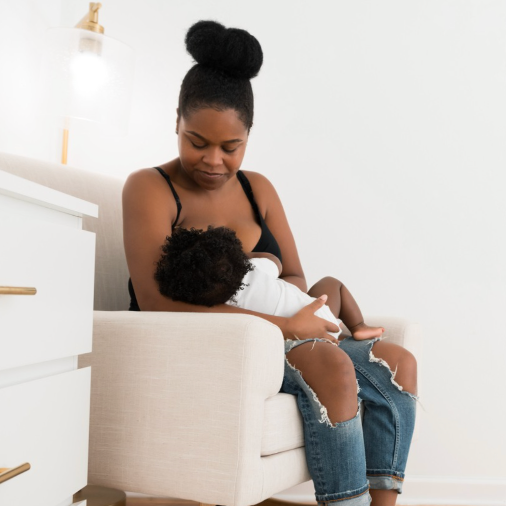 A woman in a black tank top and ripped jeans breastfeeds a baby sitting on her lap in a bright, minimalist room.