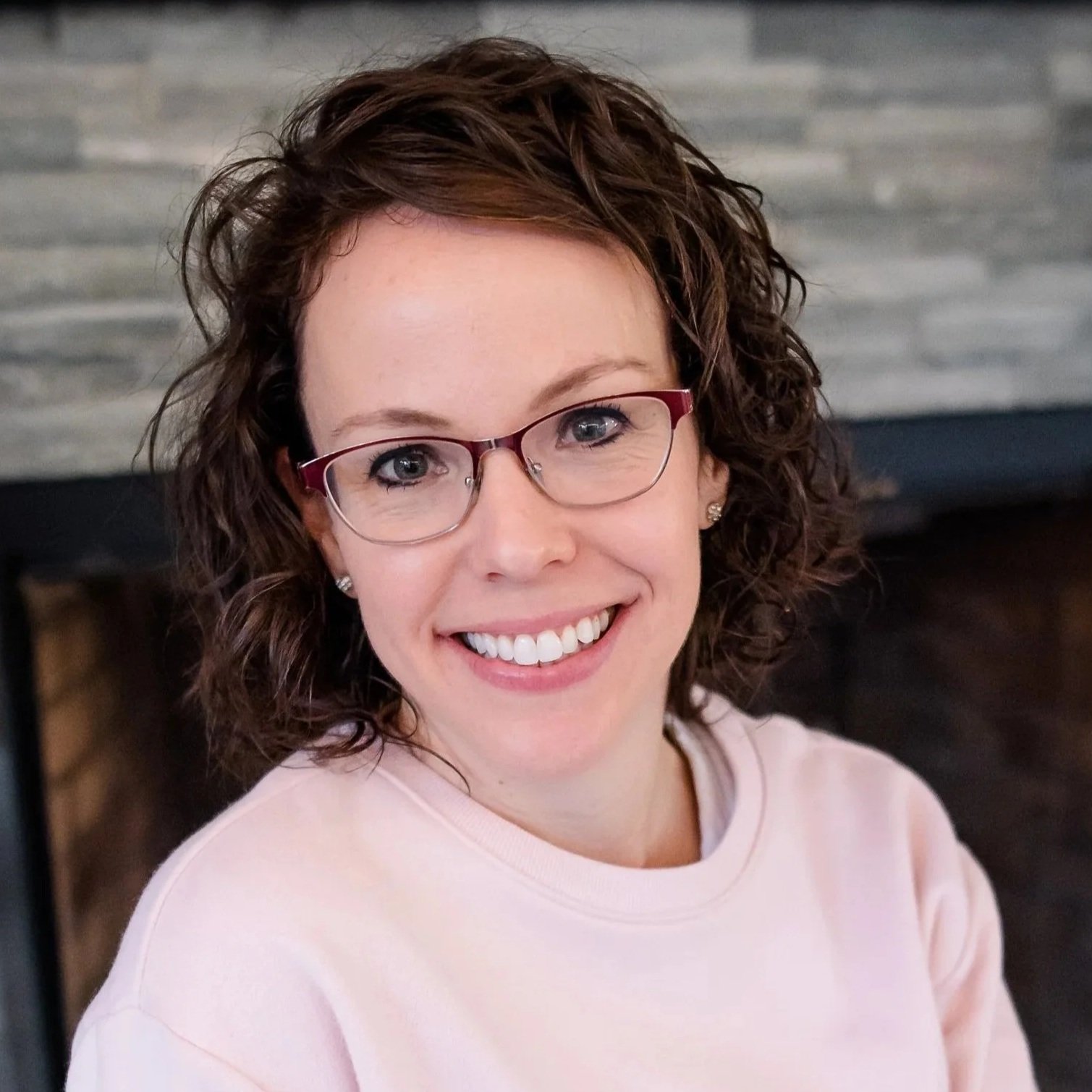 A woman with curly brown hair, glasses, and a pink shirt smiling at the camera inside a room with a stone wall background.