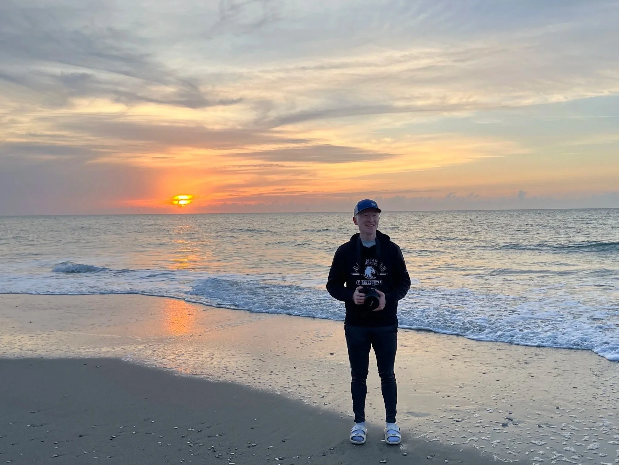A young man standing on a beach at sunset, holding a camera, with the ocean and a colorful sky in the background.