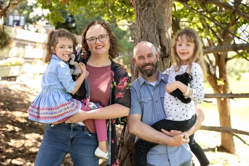 Family of four smiling beneath a leafy tree beside a wooden fence during an outdoor portrait session