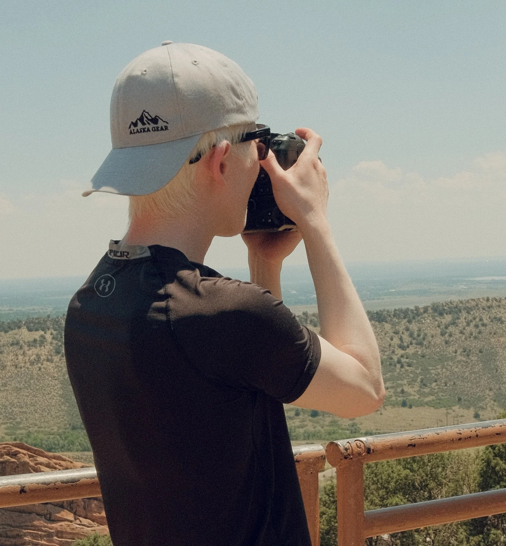 A person with blonde hair, wearing a gray baseball cap with an Alaska Gear logo, sunglasses, and a black shirt, taking a photo with a camera in an outdoor setting with a landscape, trees, and sky in the background.