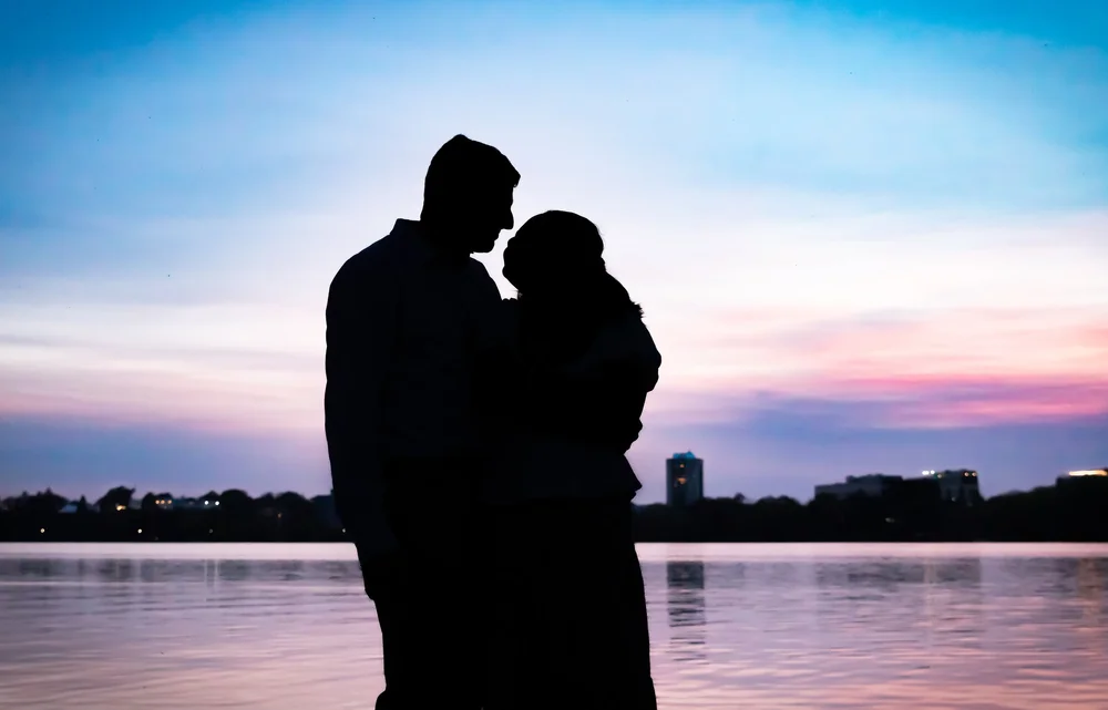 Silhouette of a couple embracing beside a lake under a pink and blue sunset sky