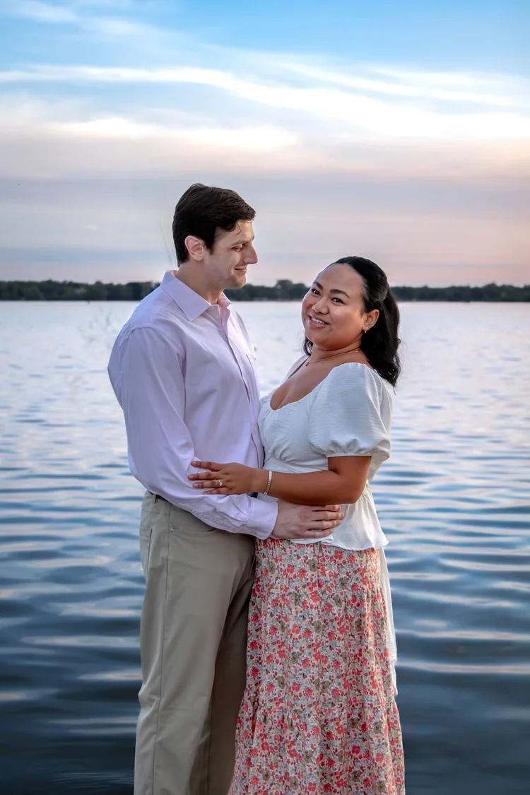 Couple holding each other beside a calm lake during a softly lit sunset portrait session