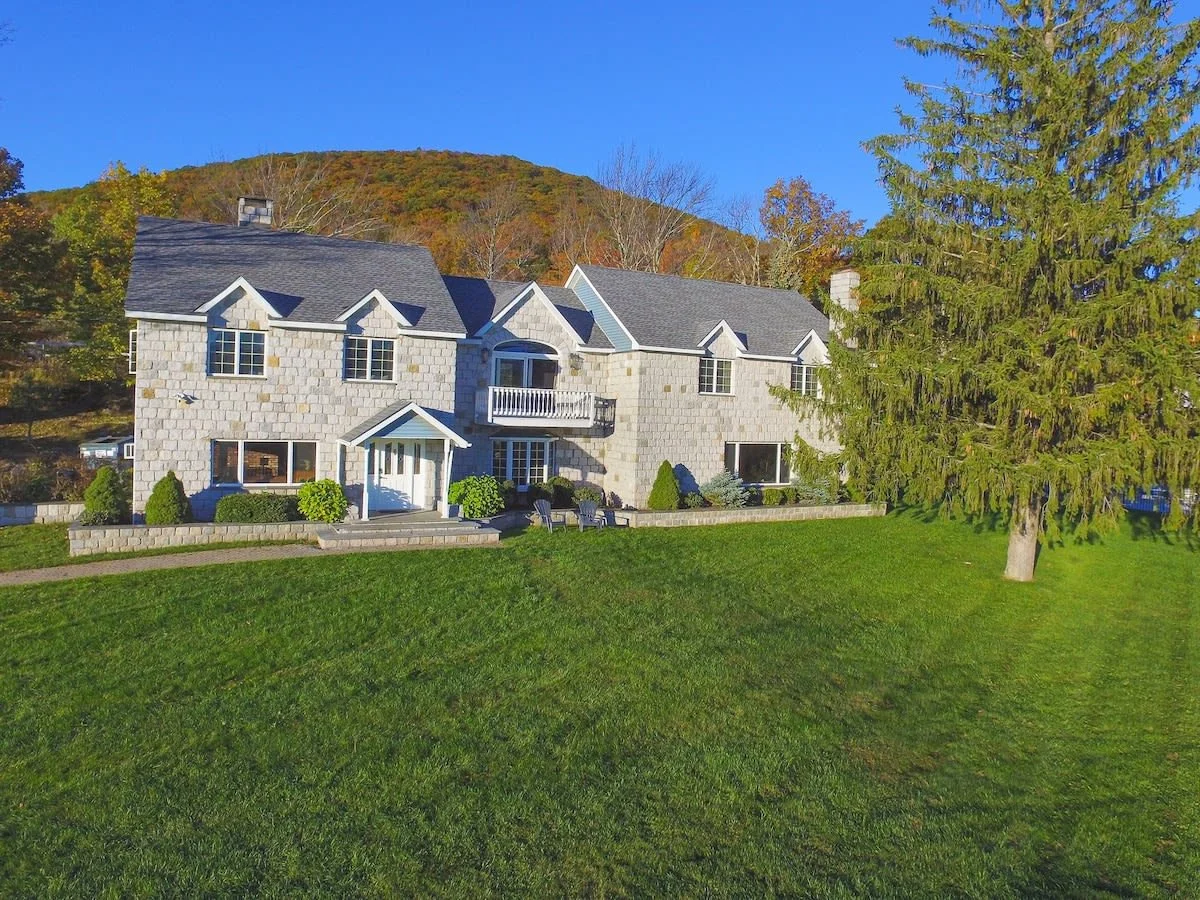 Stone rental house with gray roof, large lawn, tall tree, and wooded mountain in the background under a clear blue sky.