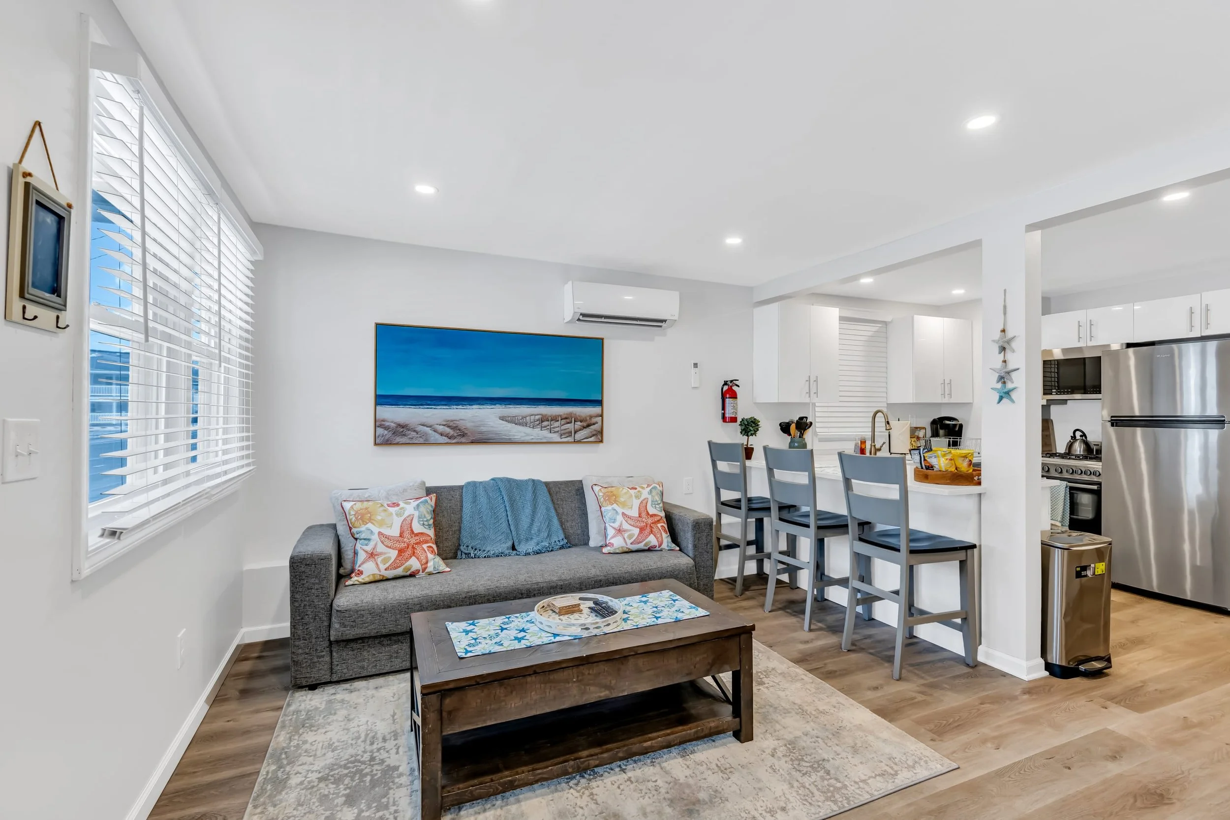 Living room of a rental house with a gray sofa, colorful starfish pillows, a wooden coffee table on an area rug, a wall-mounted TV, a blue ocean photo, and a view into a white kitchen with bar stools and small appliances.