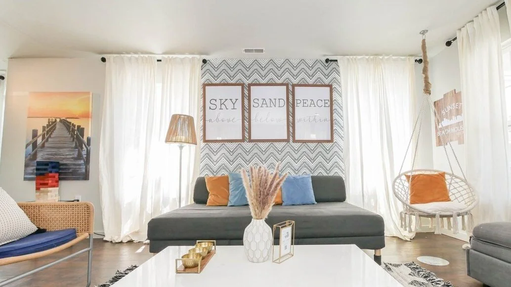 Cozy living room in a rental house with white curtains, a dark gray sofa with blue and orange pillows, a hanging chair with an orange pillow, a framed picture of a pier at sunset, and wall art with sky, sand, and peace themes.