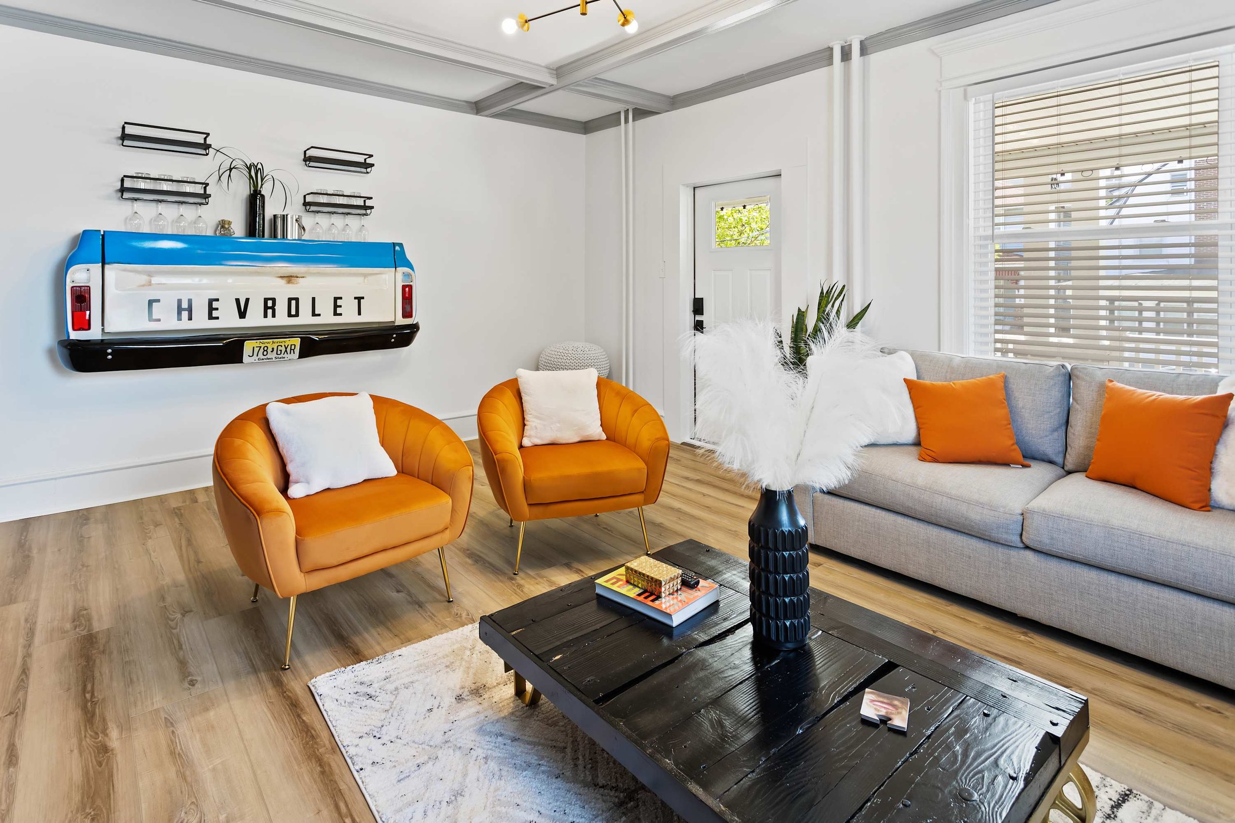 Living room featuring a gray couch with orange pillows, two orange armchairs with white pillows, a black wooden coffee table with a vase of white feathers, a chevrolet truck tailgate decor on the wall with shelves, a door, large windows with blinds, and a light-colored rug on wooden floors.