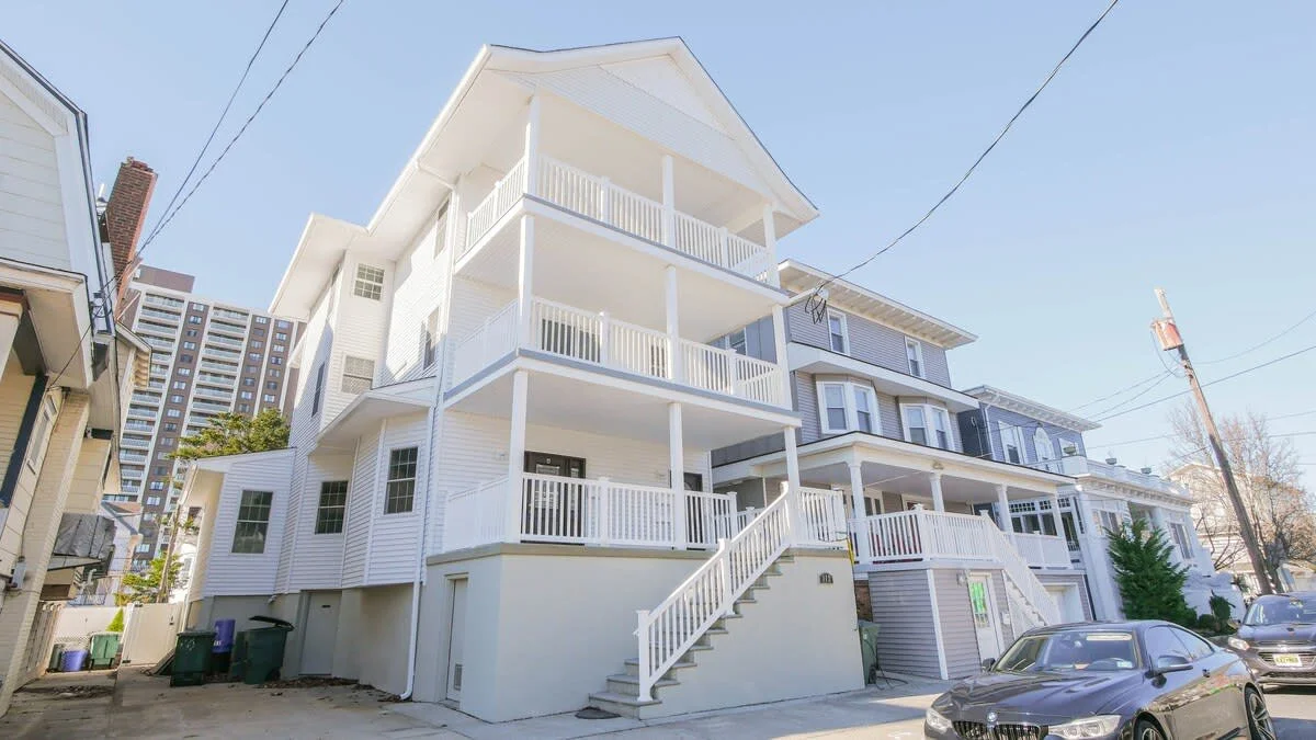 Multi-story residential rental building with white and gray exterior, multiple balconies, and a staircase leading to the entrance, cars parked in front, clear blue sky