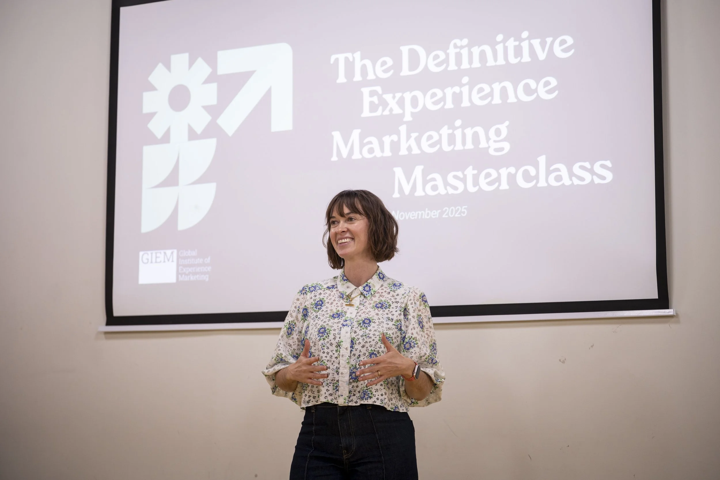 A woman with shoulder-length brown hair, wearing a floral blouse, speaking in front of a large screen that displays the words 'The Definitive Experience Marketing Masterclass' and the date 'November 2025'. She appears to be giving a presentation or lecture.