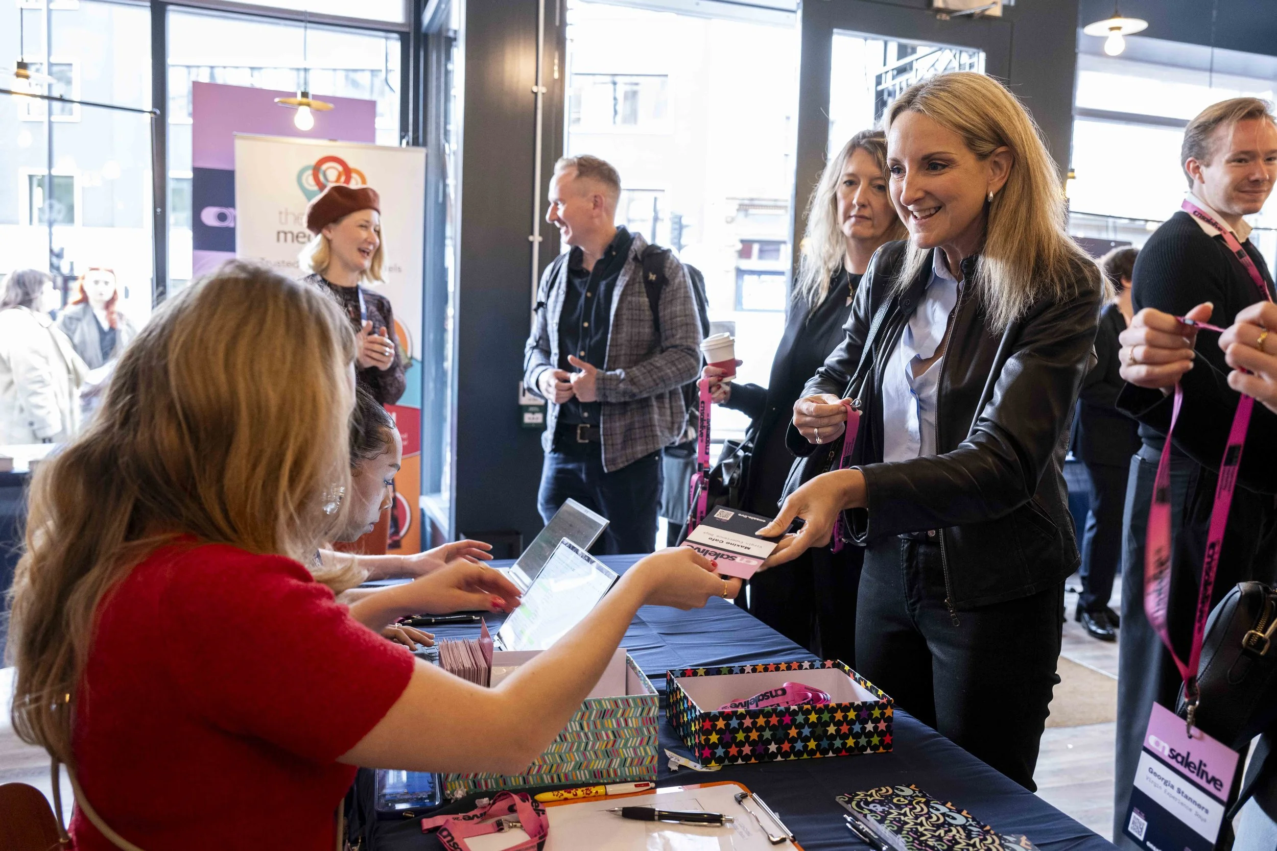 At a conference registration table, a woman with blonde hair wearing a black leather jacket is smiling while receiving a badge or lanyard from a woman in a red sweater. Several people are in the background engaging in conversations, and there are reg