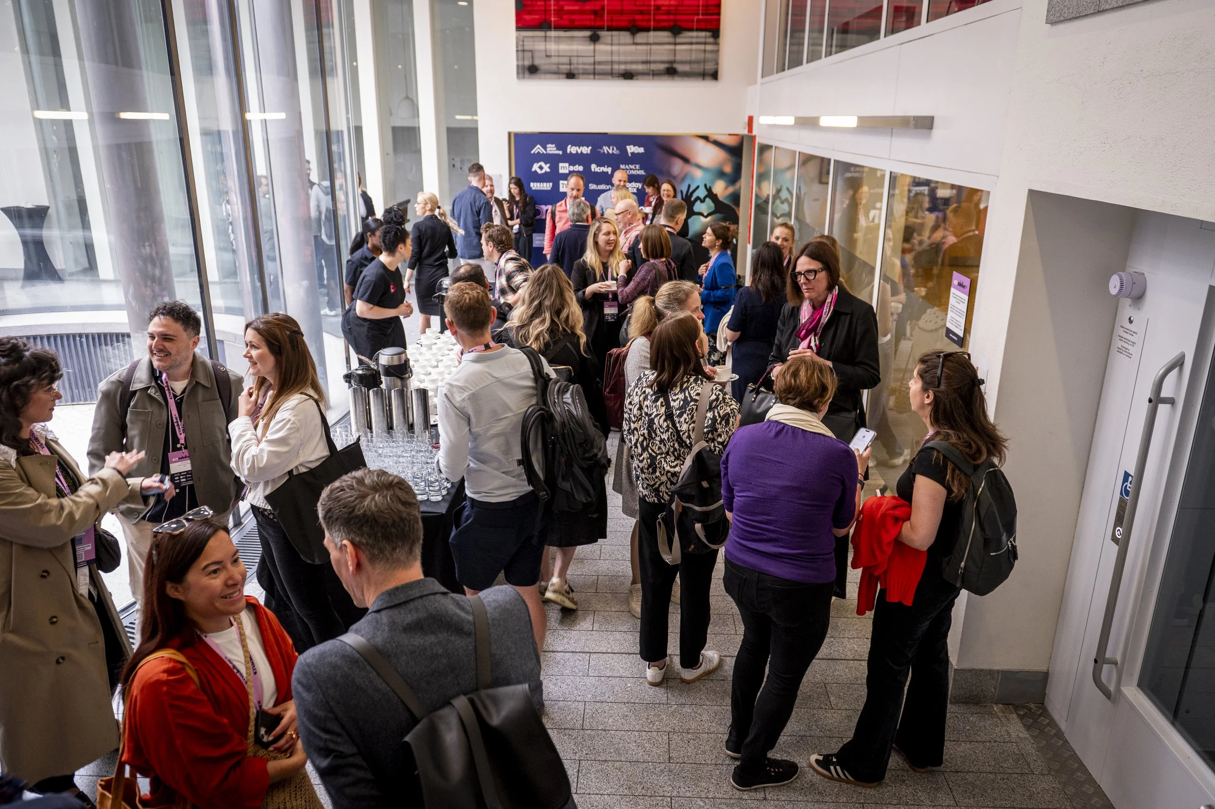 People gathered at a busy conference registration or check-in area, some talking and others using smartphones, with a refreshment table with water bottles on the left side, and a large digital screen and glass windows in the background.