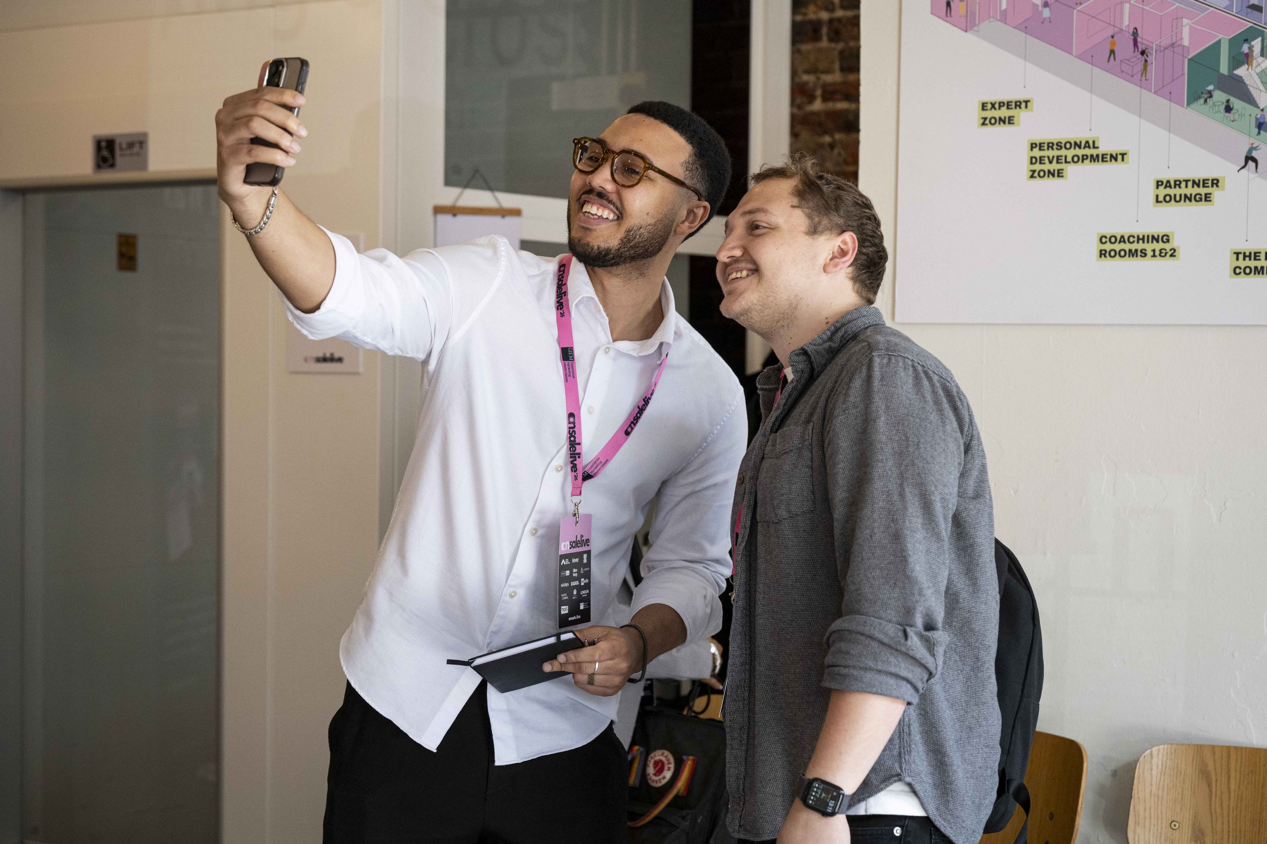 Two men are taking a selfie inside a building with one man holding a smartphone. One man is wearing glasses and a white shirt, and the other is wearing a gray shirt and a smartwatch. Behind them is a wall with a colorful map or diagram with labeled z