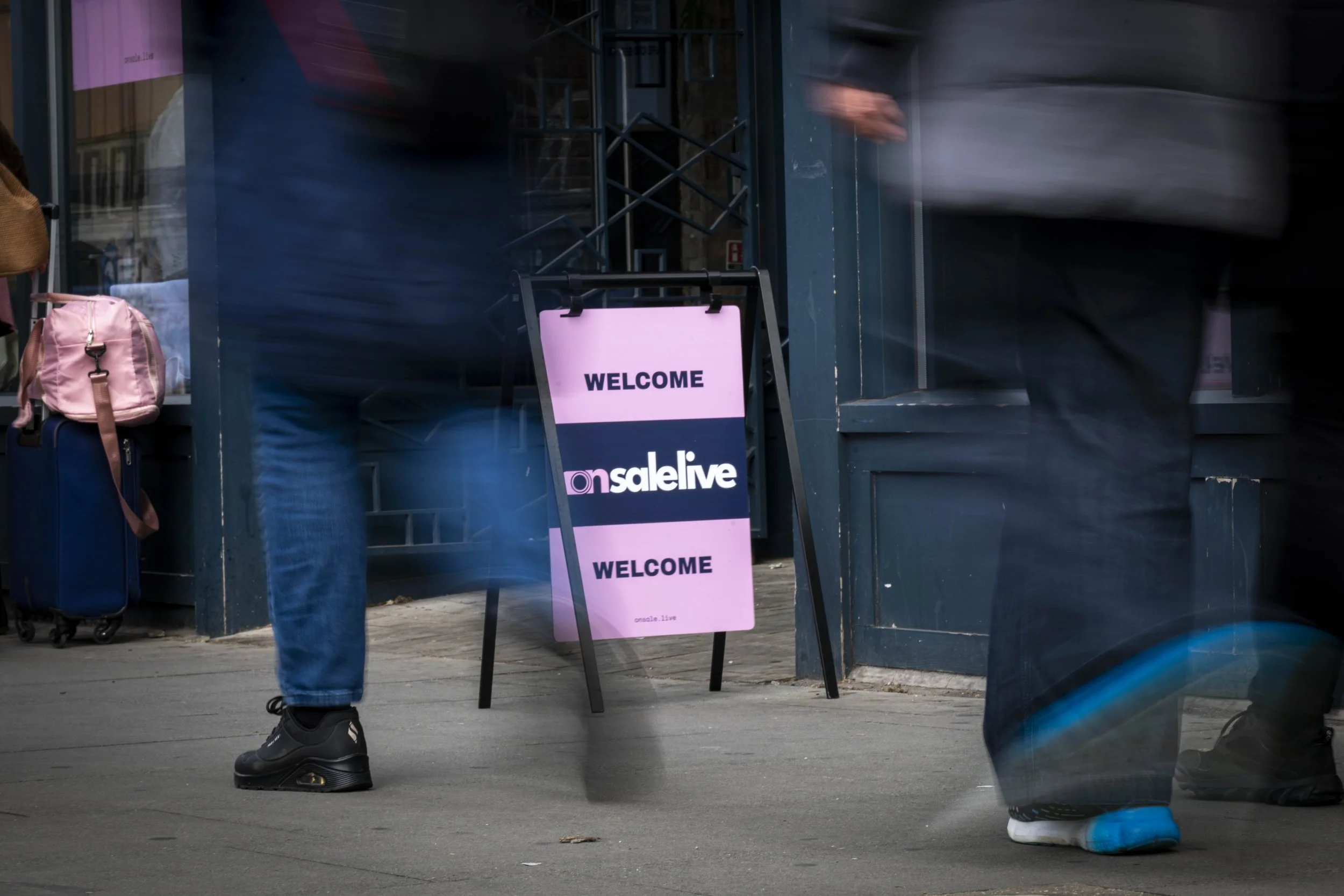 People walking past a storefront entrance with a pink sign that reads 'WELCOME' and a black sign below that reads 'onsale.live.' There are bags and luggage near the entrance, and the photo captures the lower half of the pedestrians, including their s