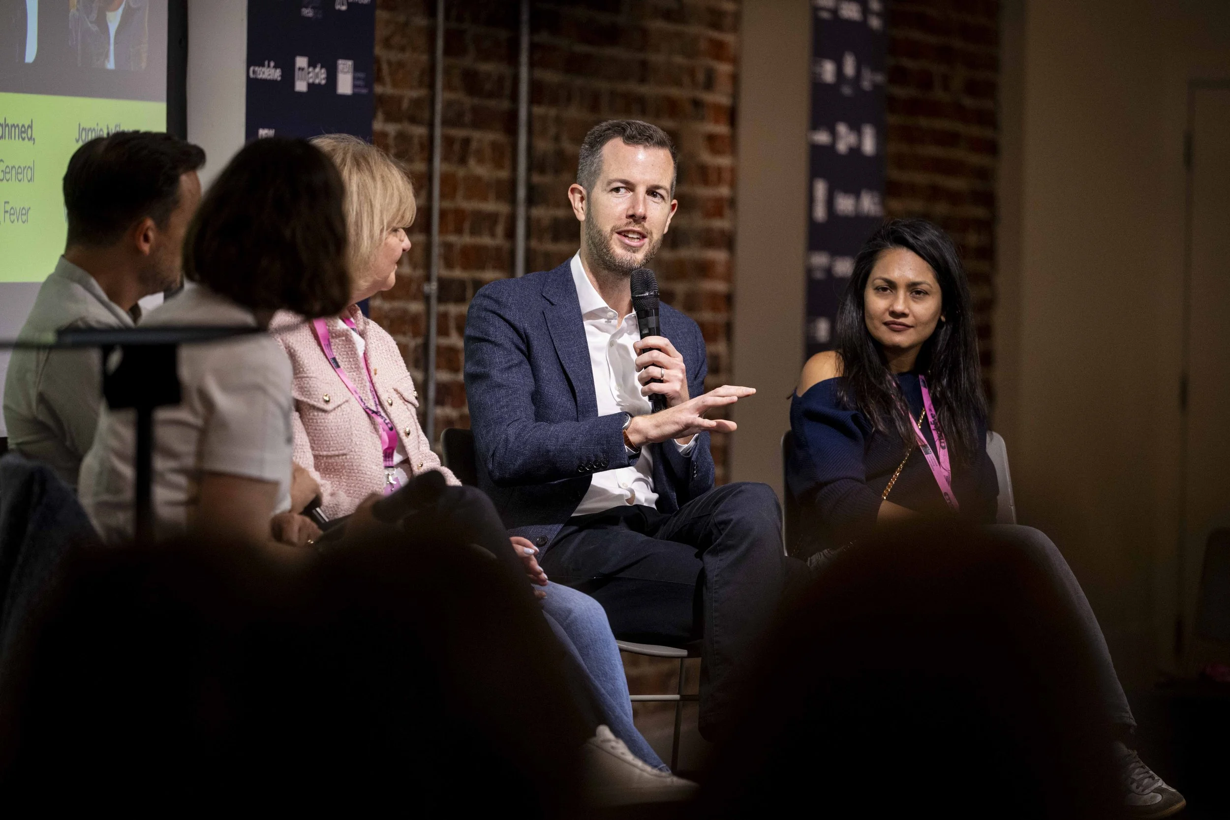 A panel discussion with five diverse individuals seated on stage, one man speaking into a microphone while others listen, set against a brick wall with event banners.