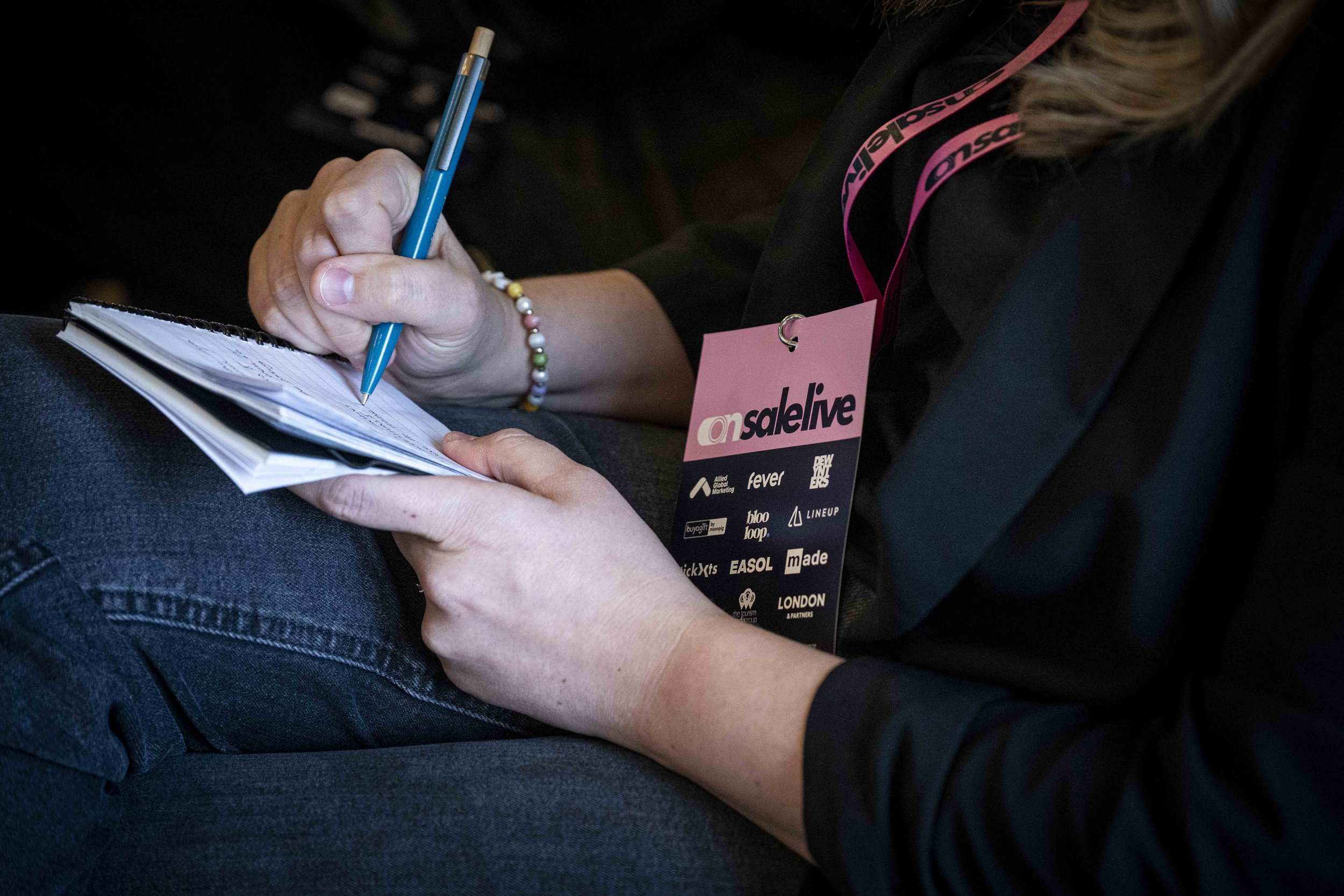 Close-up of a person holding a blue pen and writing in a notebook, wearing a black shirt with a pink strap and a colorful beaded bracelet, seated with crossed legs.