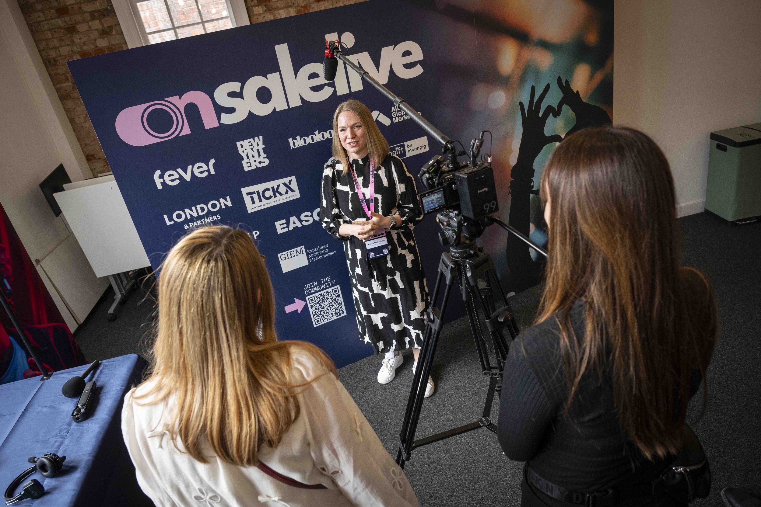 A woman speaking at a conference or event with a large backlit sign behind her that reads 'on sale live' and includes logos for various brands. She is being filmed by a camera operator.