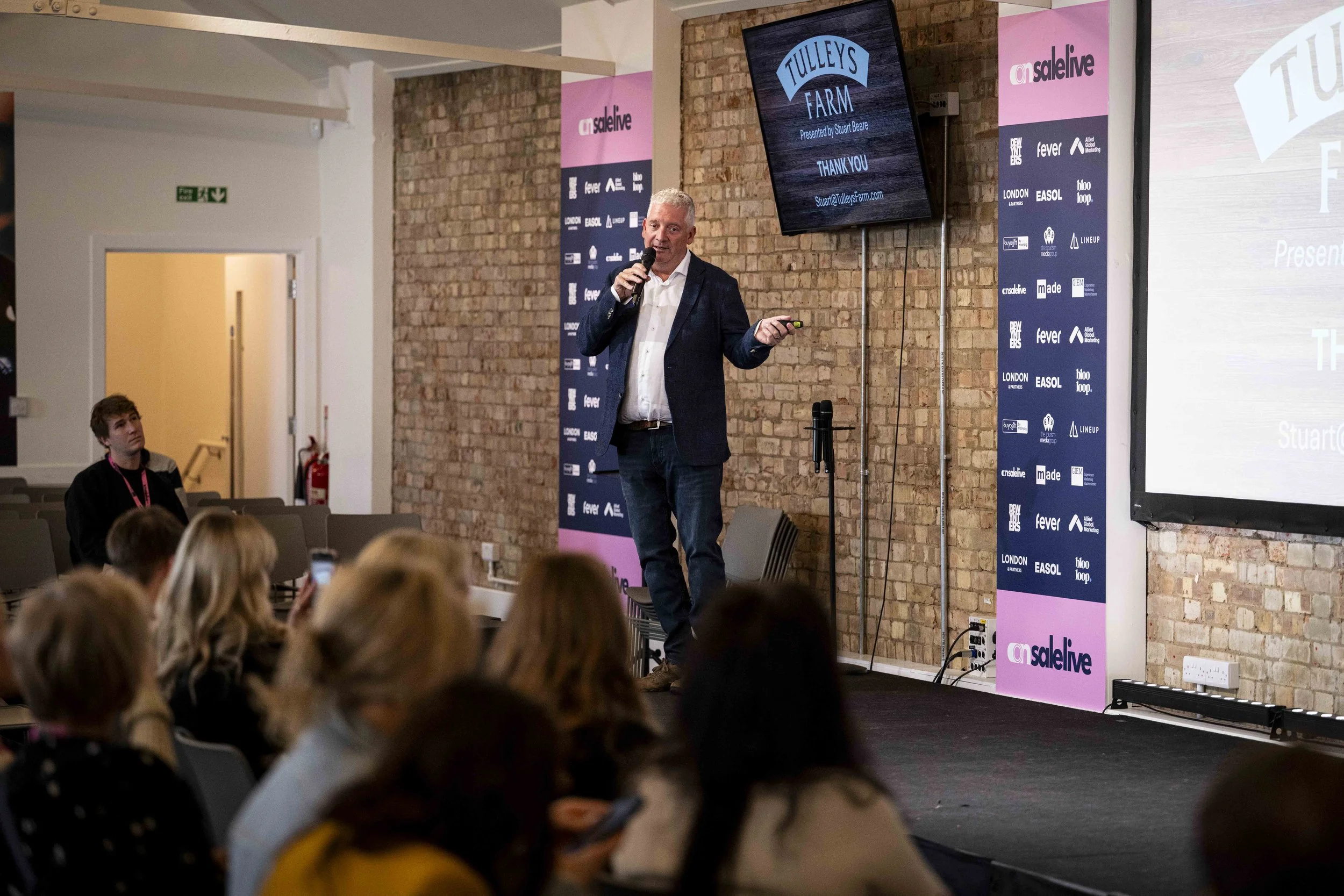 A man in a dark blazer and white shirt giving a presentation with a microphone in hand in front of a brick wall with digital screens and banners in a conference room filled with seated audience members.