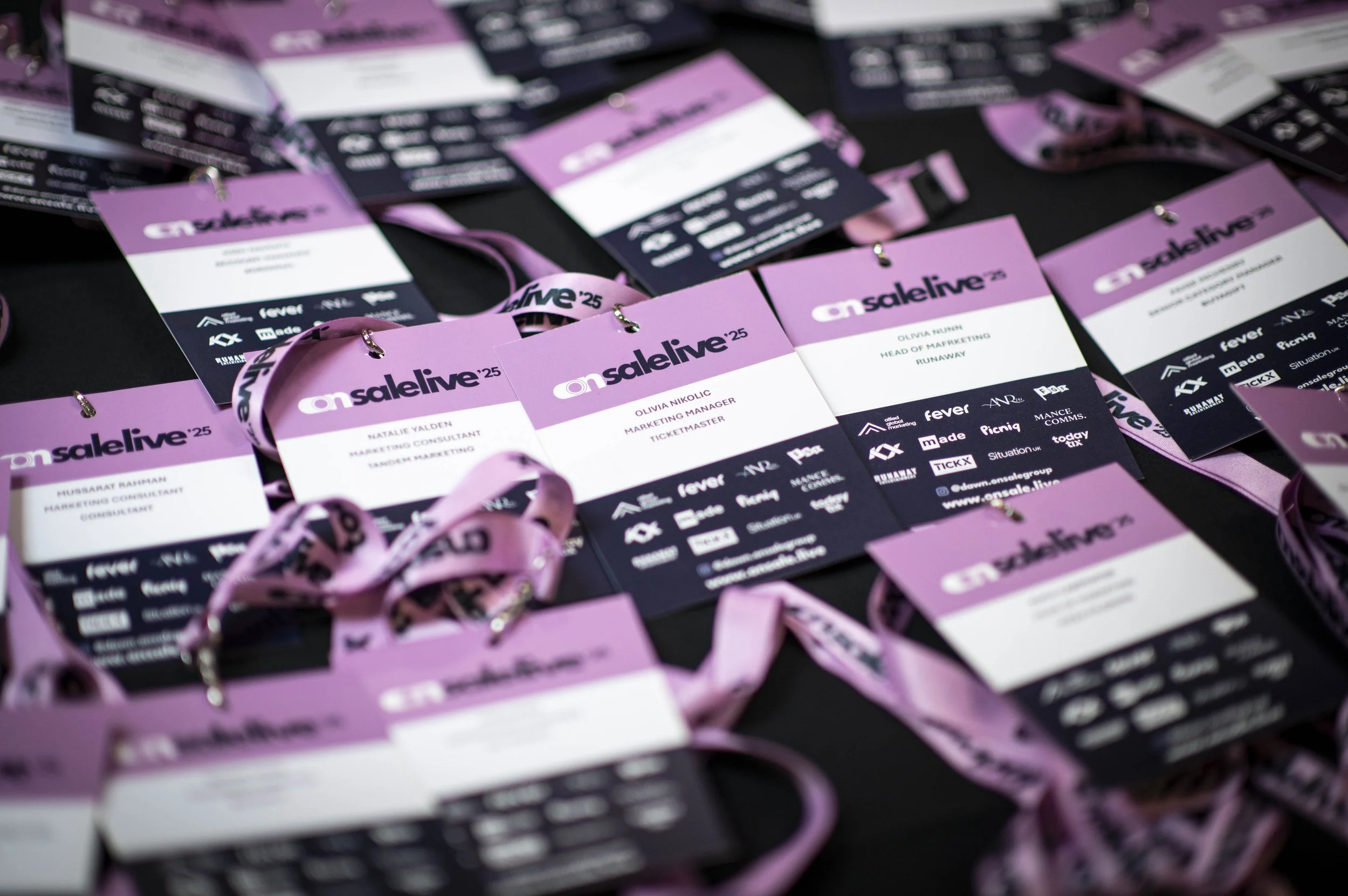 Conference badges for an event called 'onsalive' with pink lanyards, spread out on a black surface, featuring attendee names and titles.