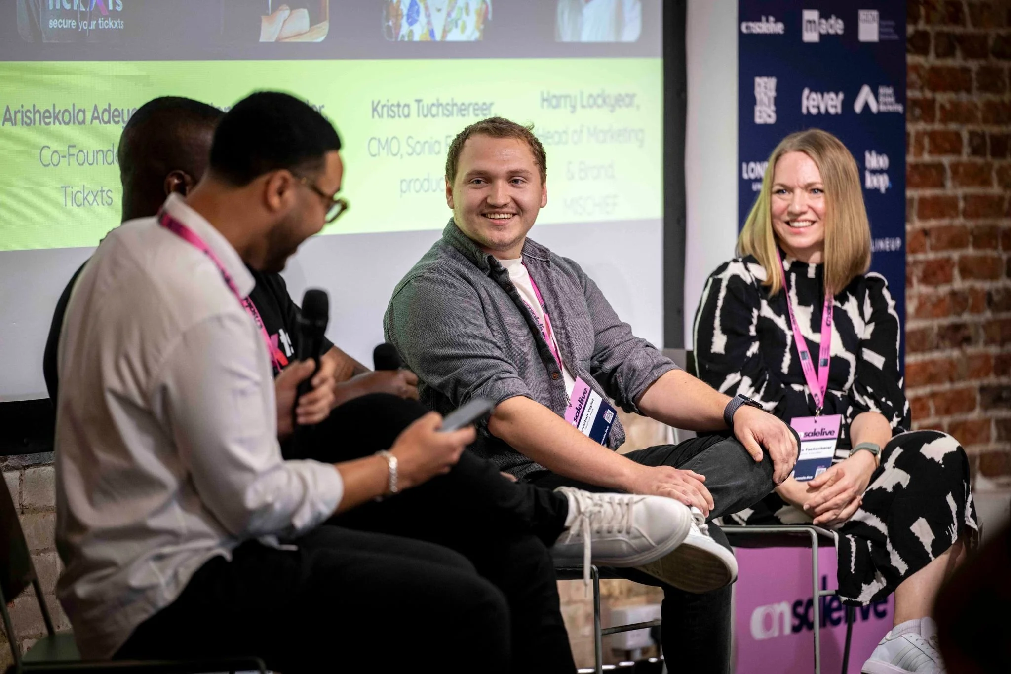 Four people sitting on chairs participating in a panel discussion at an event with a brick wall background and a large screen behind them displaying text and logos.