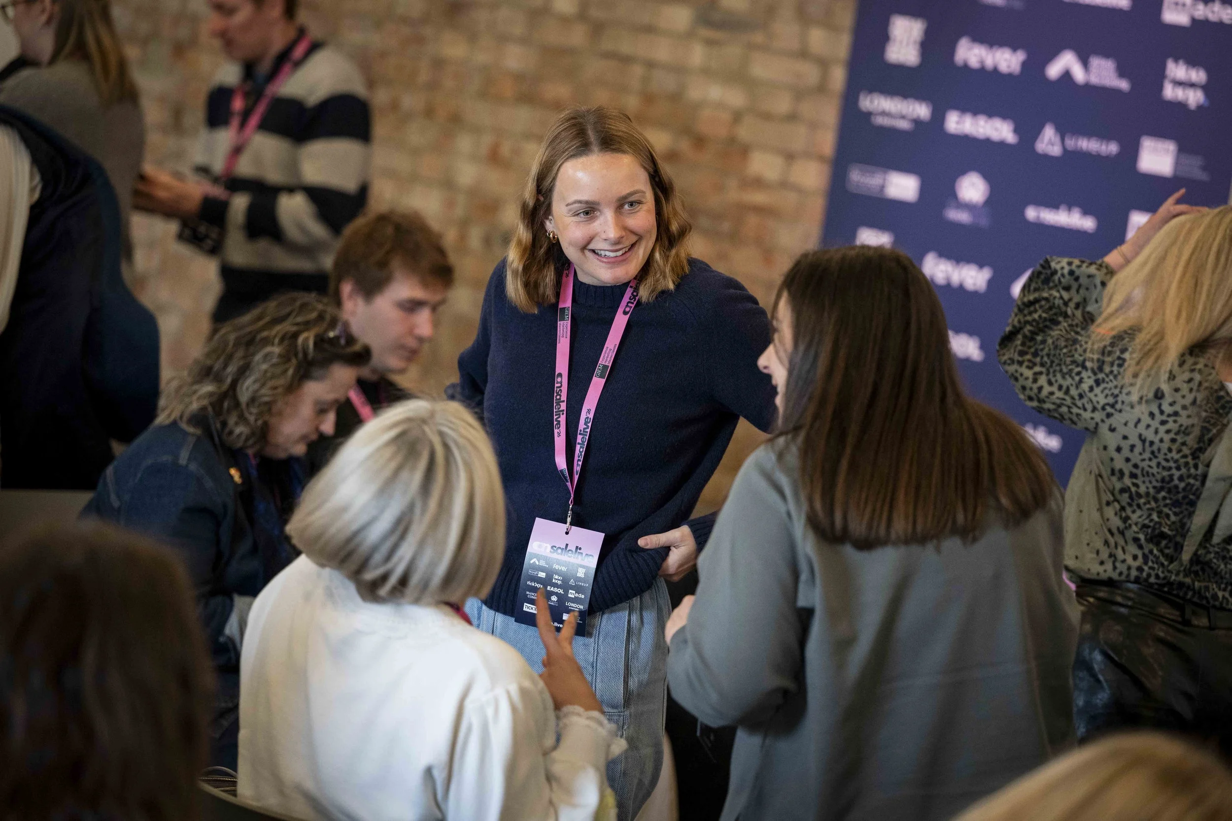 A smiling woman with shoulder-length brown hair talking to a group of women at a conference or event, with a blue branded banner in the background and people wearing event lanyards.