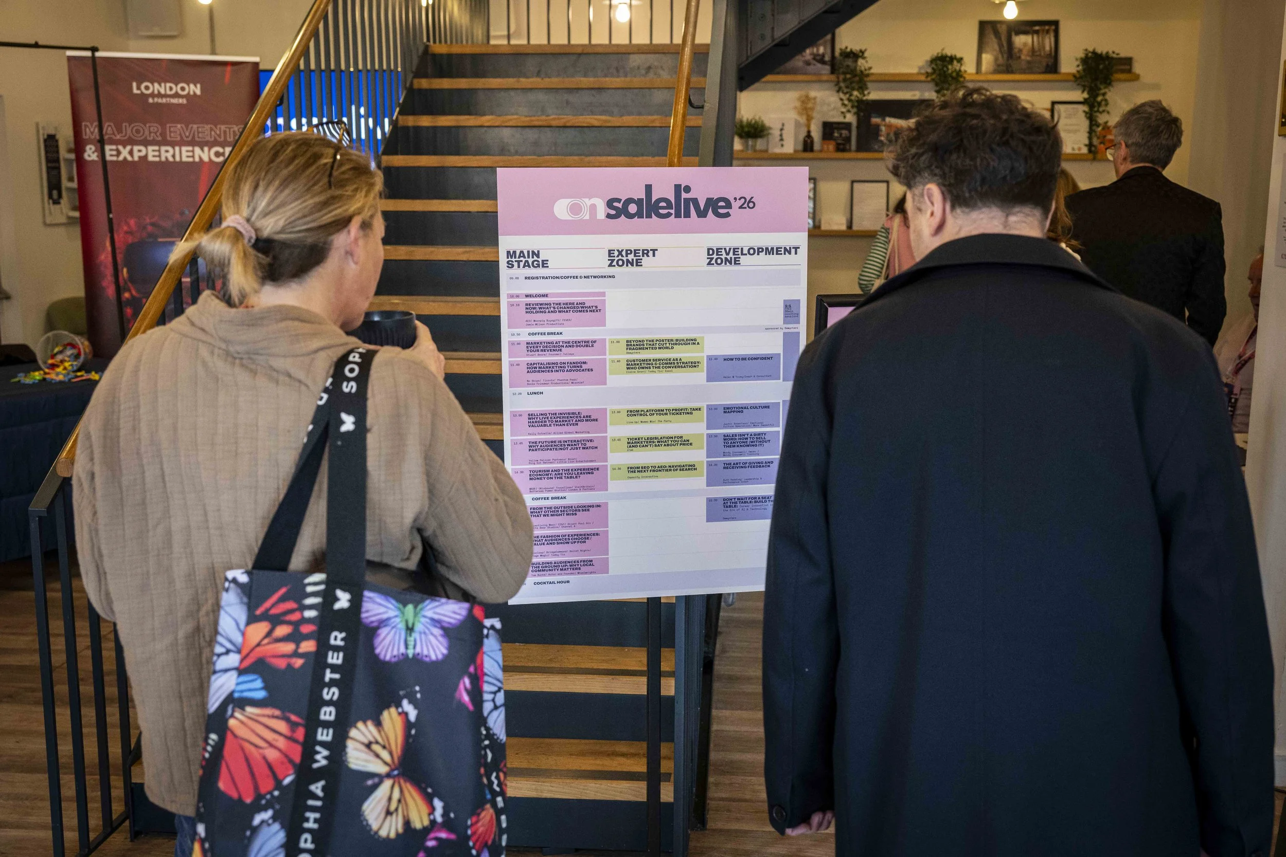Conference attendees, including a woman with a beige shirt and a man with dark curly hair, standing in front of a schedule board at the 'Salelive '26' event. The schedule lists events on different zones, under a main stage, expert zone, and developme
