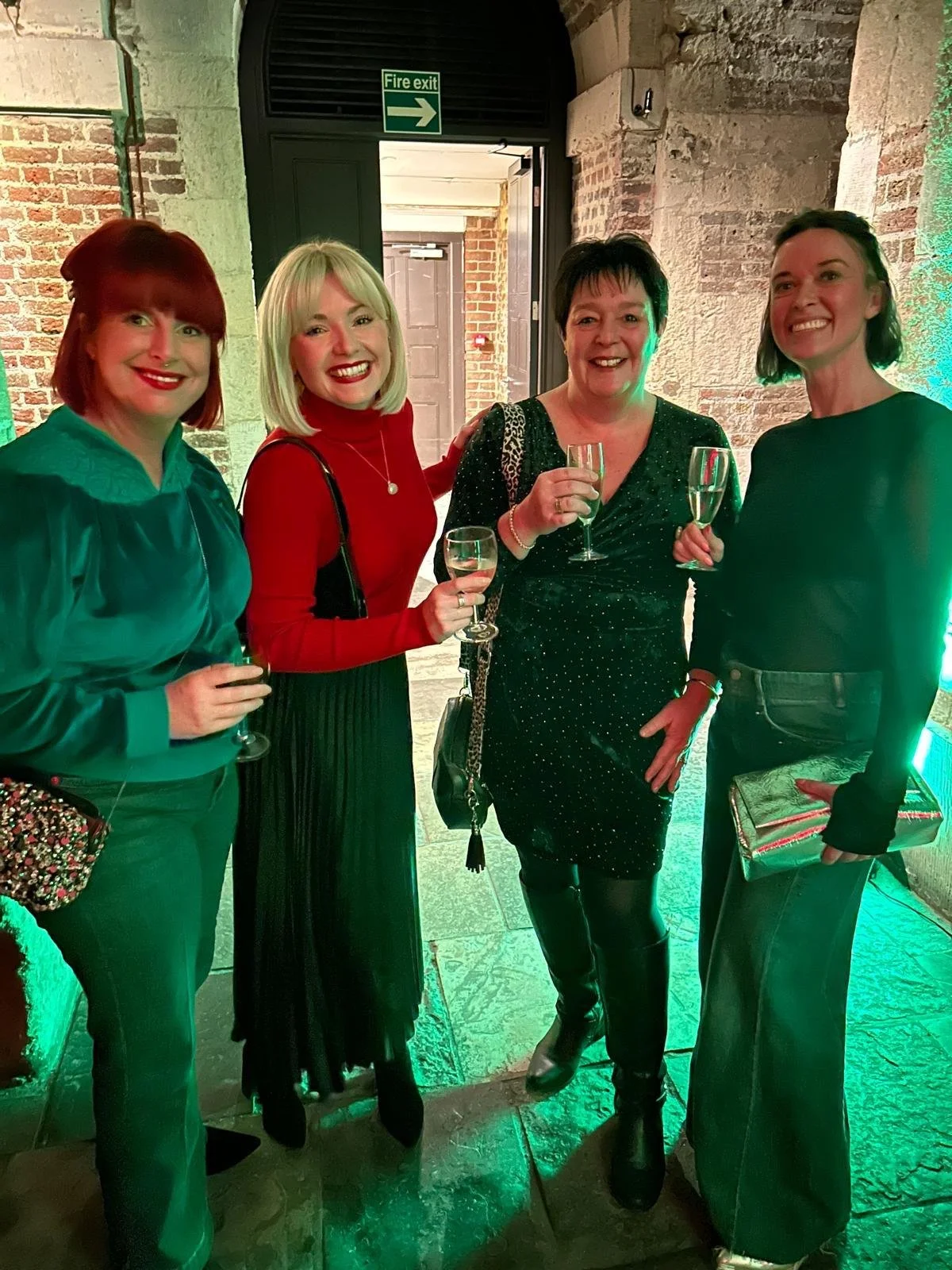 Four women standing together at a celebration, holding glasses of champagne, inside a brick-walled venue with green lighting.