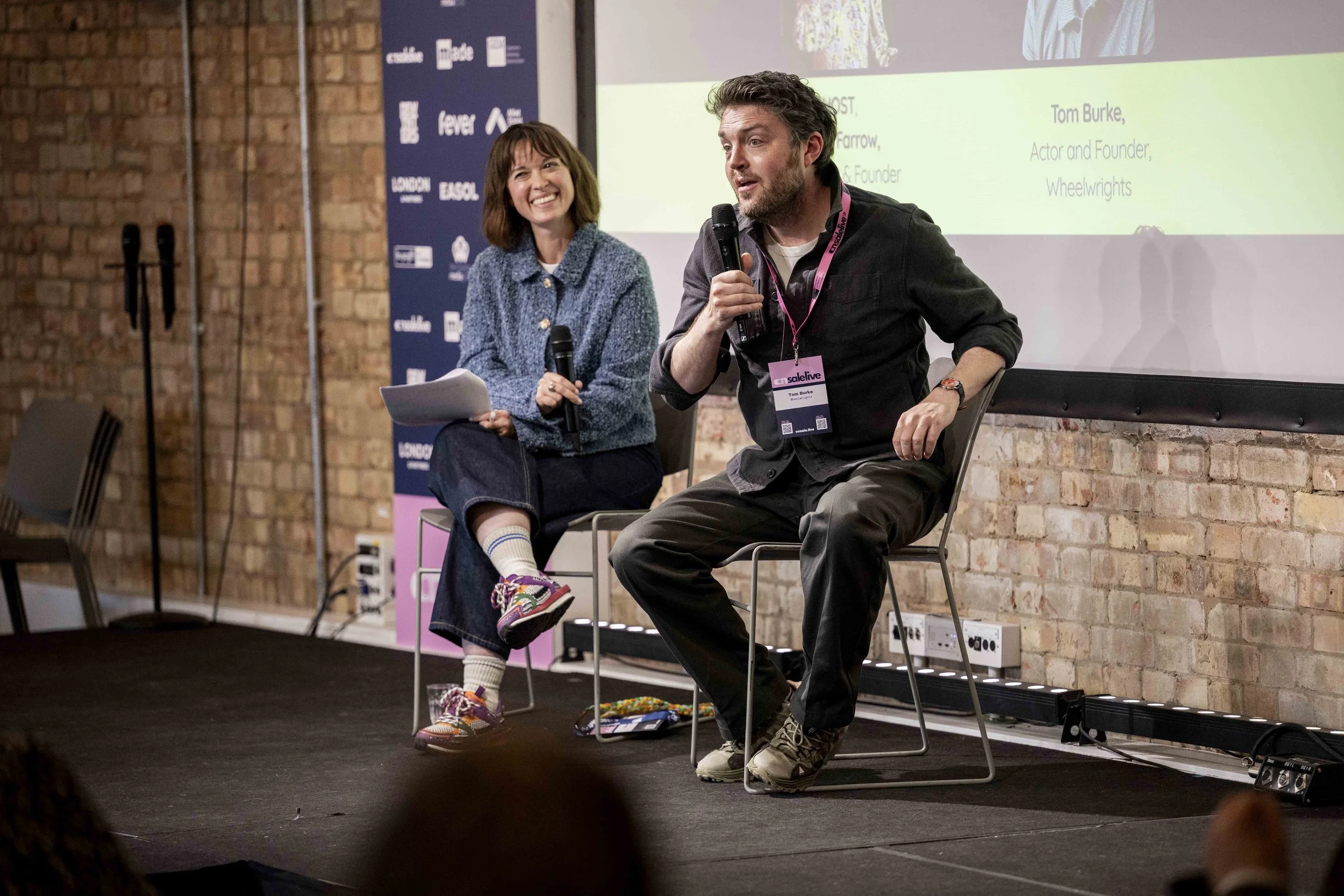 A man and woman seated on stage during a discussion or panel at an event, speaking into microphones. The woman is smiling, wearing a blue sweater, and holding papers. The man has a beard, wearing a black shirt with a lanyard and name badge, and is sp