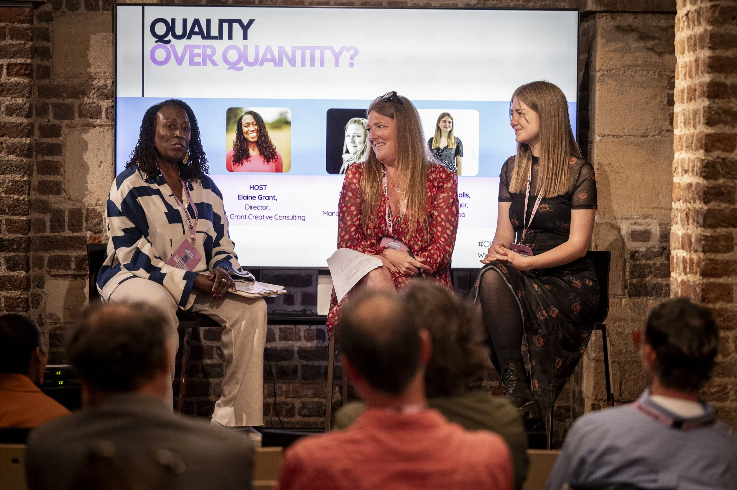 Three women sitting on chairs engaged in a discussion on stage at a conference or panel event. Behind them is a large screen displaying the text 'Quality Over Quantity?' and images of women, with their names and titles. Audience members are seated in front.