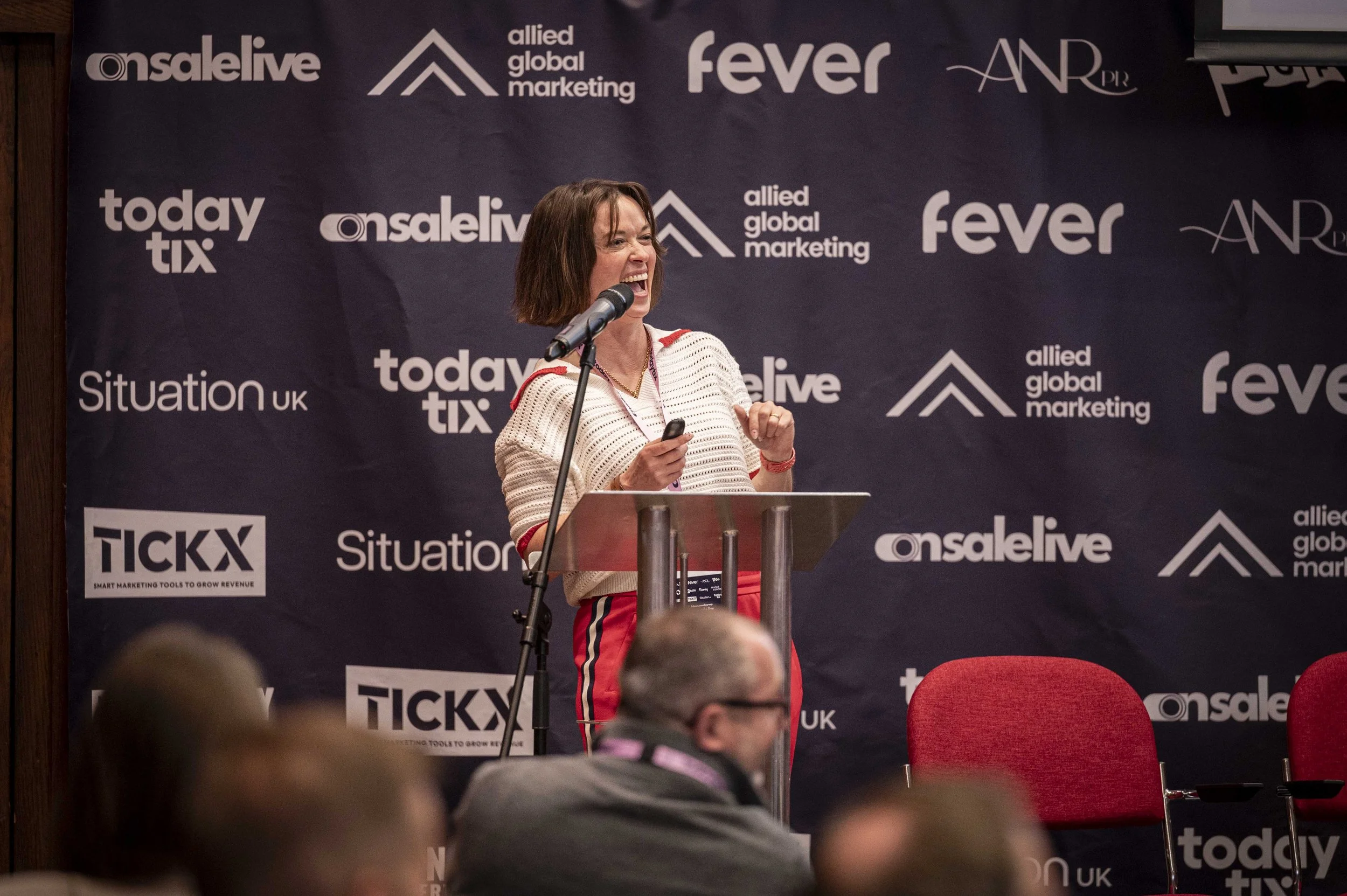 A woman speaking passionately at a podium during a conference or event, with a dark backdrop featuring various sponsor logos and text.