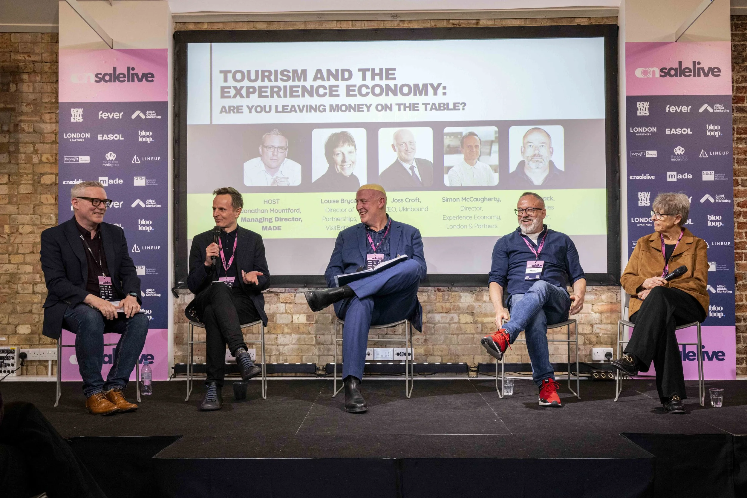 Panel discussion at a conference with five panelists sitting on stage, a large screen behind them displaying the panel topic 'Tourism and the Experience Economy: Are You Leaving Money on the Table?' and photos of the speakers, brick wall background, 