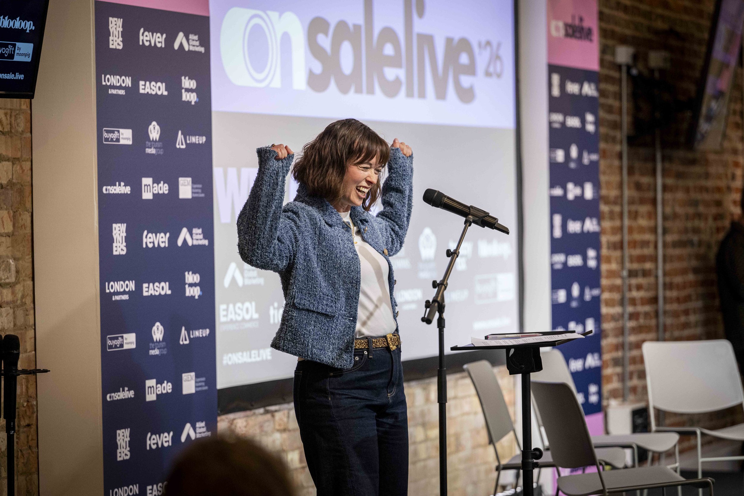 A woman standing at a podium, smiling and raising her arms in celebration, in front of a large screen and a backdrop with various sponsor logos.