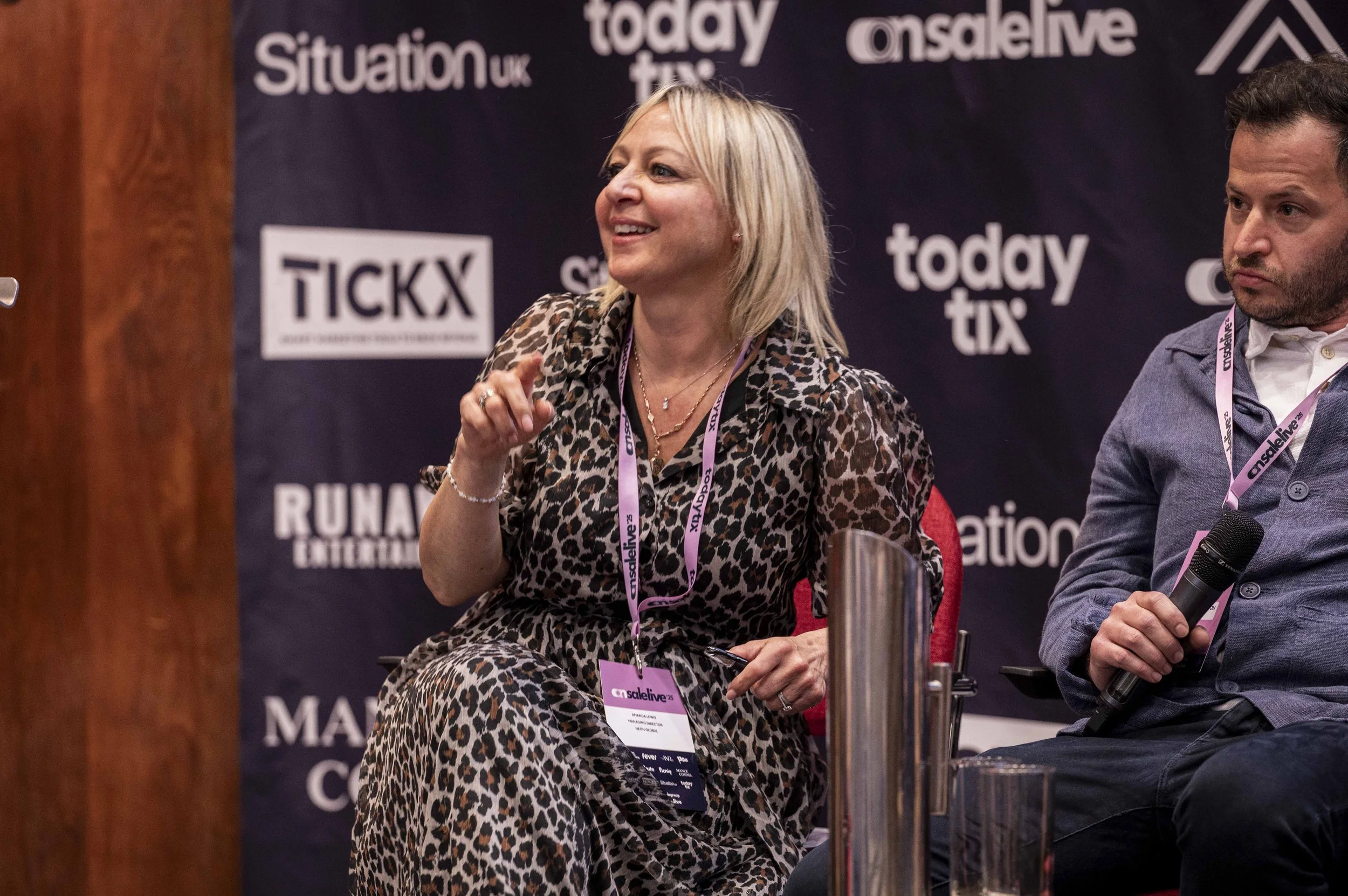 A woman with blonde hair in a leopard print dress smiling and sitting on a panel at a conference, holding a microphone, with a man next to her holding a microphone. The background features logos including 'situationuk', 'todaytix', and 'onasalive'.