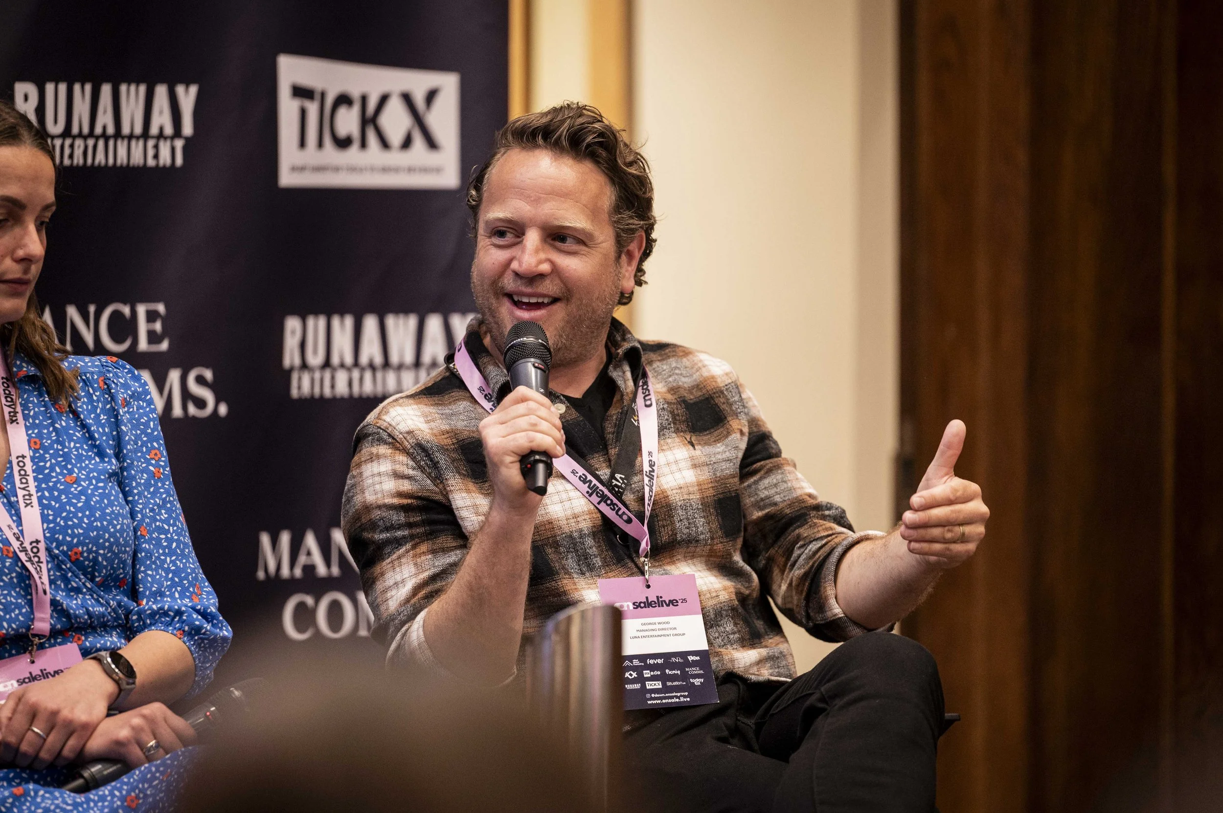 A man sitting and holding a microphone, speaking at a panel discussion, wearing a plaid shirt and a conference badge around his neck.