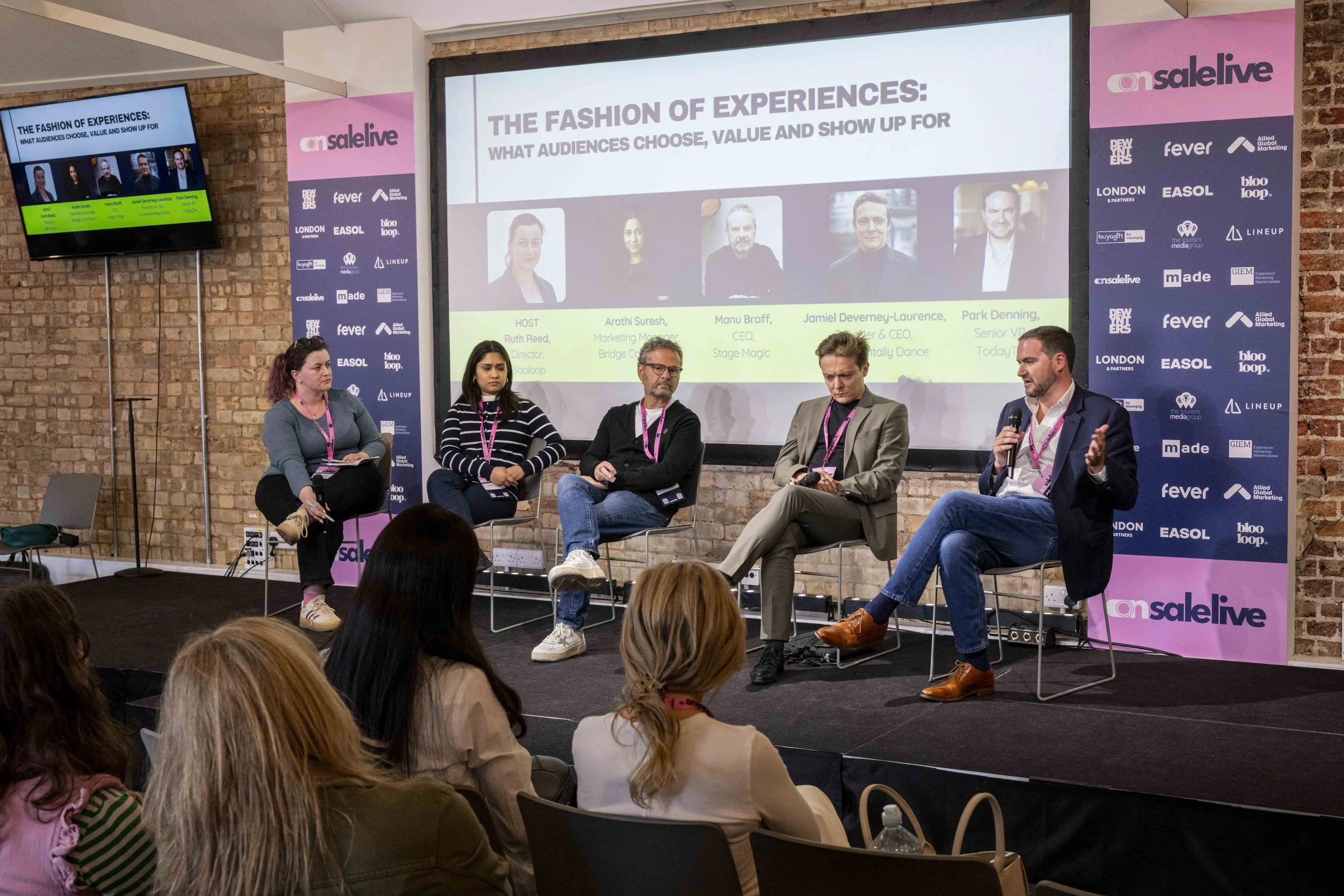 Panel discussion at a conference with five speakers on stage, audience members seated facing the stage, large screens displaying presentation slides and branding, brick wall background, and banners displaying event branding and sponsors.
