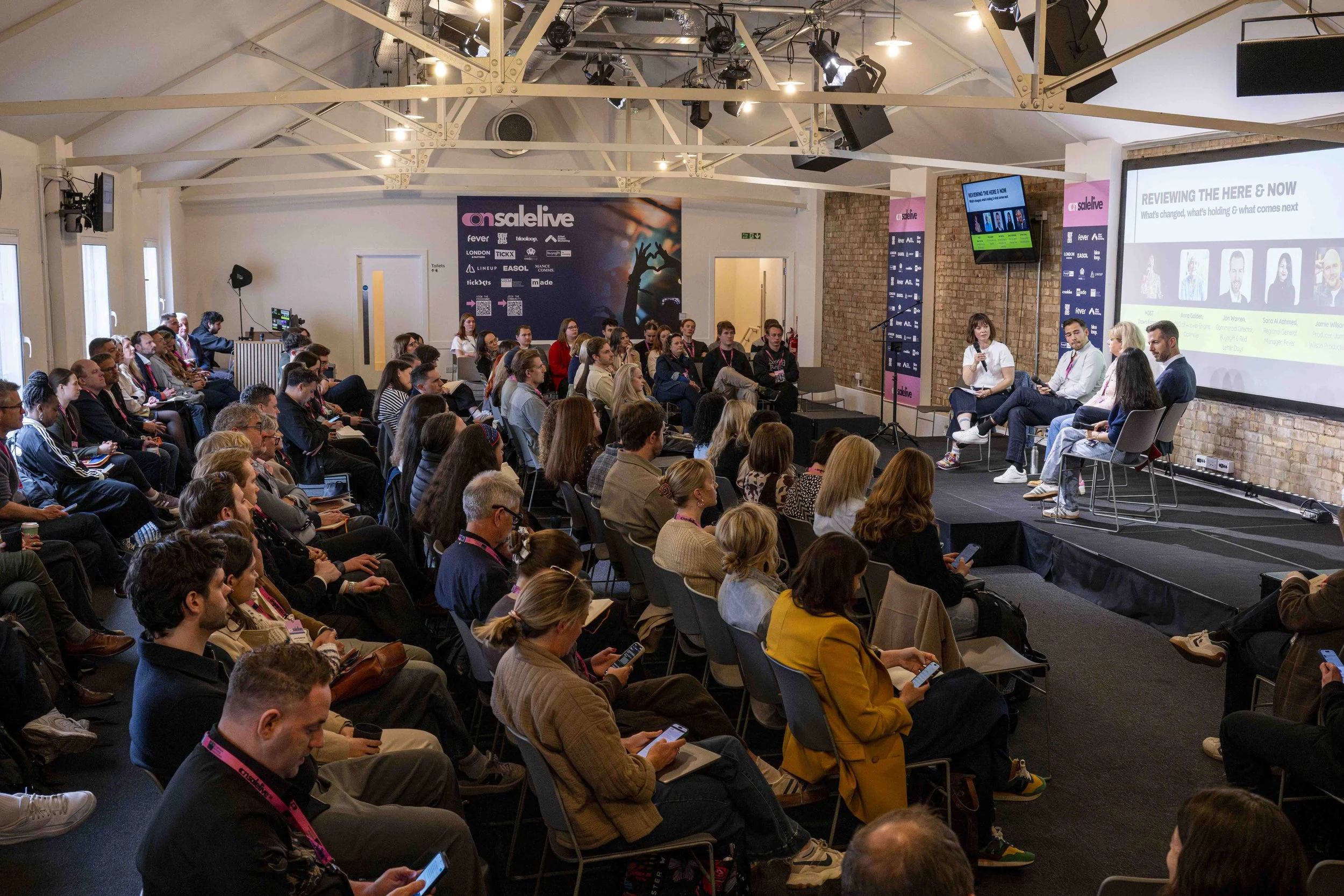 A conference room filled with seated attendees watching a panel discussion on stage. The stage has five speakers and a large screen displaying a presentation. The room has exposed brick walls, large windows, and industrial ceiling beams. The event is
