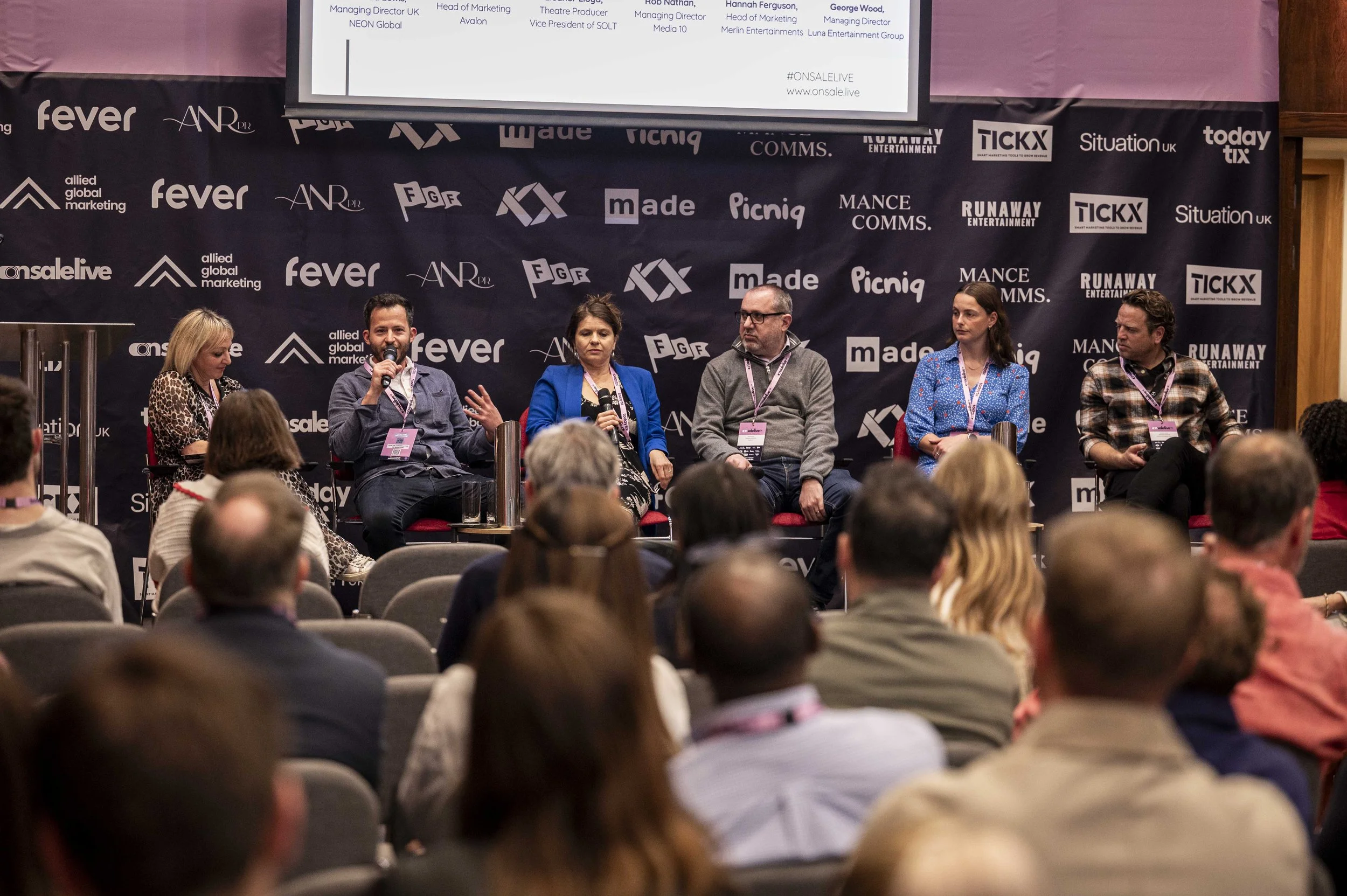 Panel discussion at a conference with five speakers seated on stage in front of a black backdrop covered with various logos. One speaker is speaking into a microphone, and the audience is seated facing the stage.