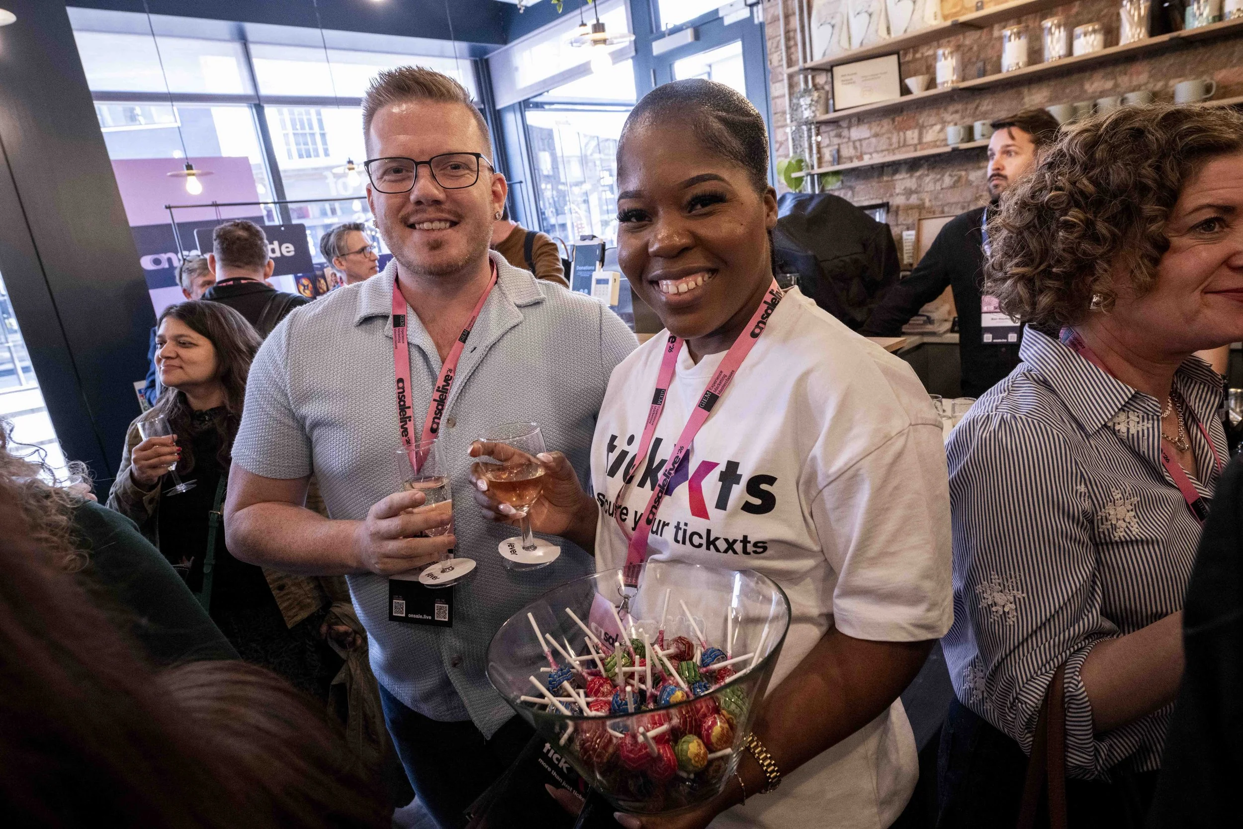 People at a social event in a cafe or restaurant, smiling and holding glasses of wine or champagne, with a bowl of assorted candies and lollipops on the table in front of them.